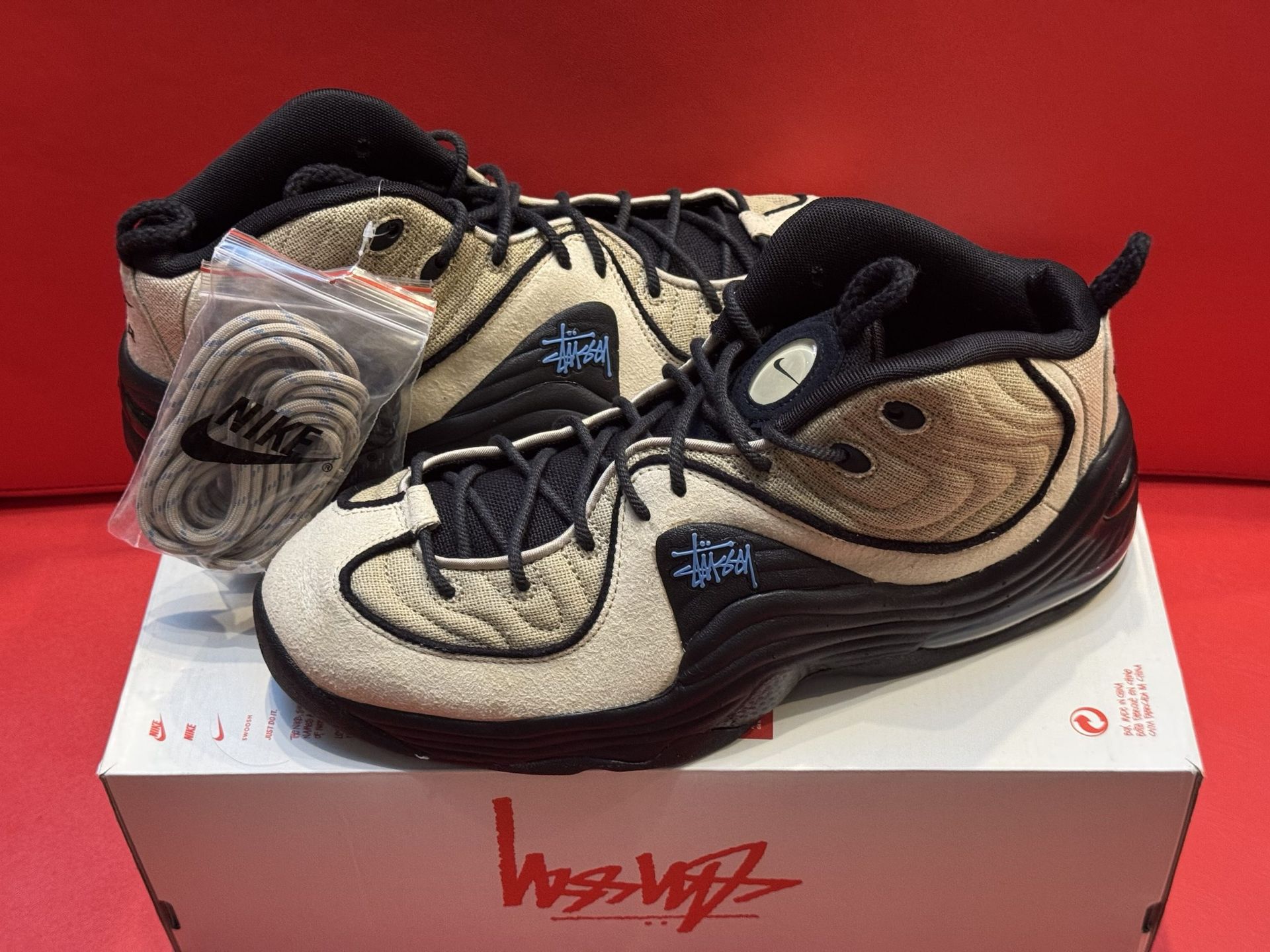 Tan and black Nike Penny Hardaway sneakers on a box with the Stussy logo, red background.