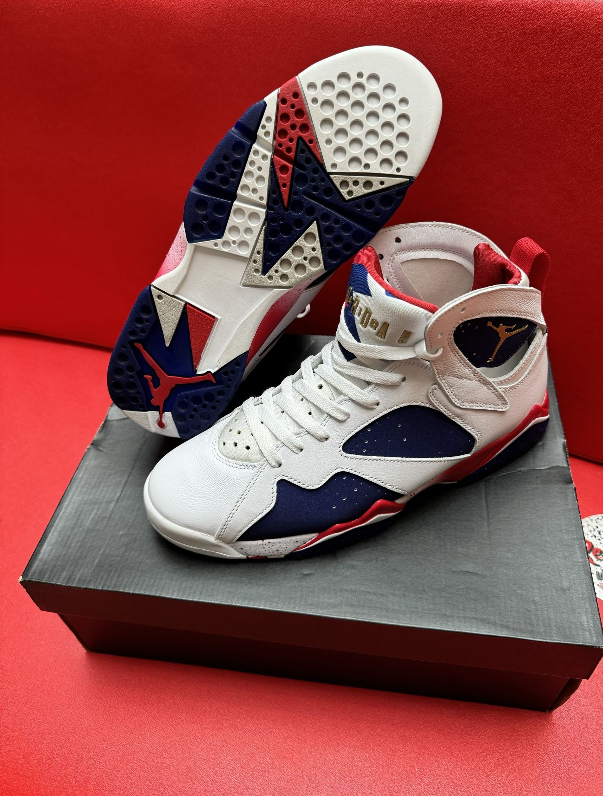 White, blue, and red Air Jordan 7 sneakers on a box, sole up, against a red background.