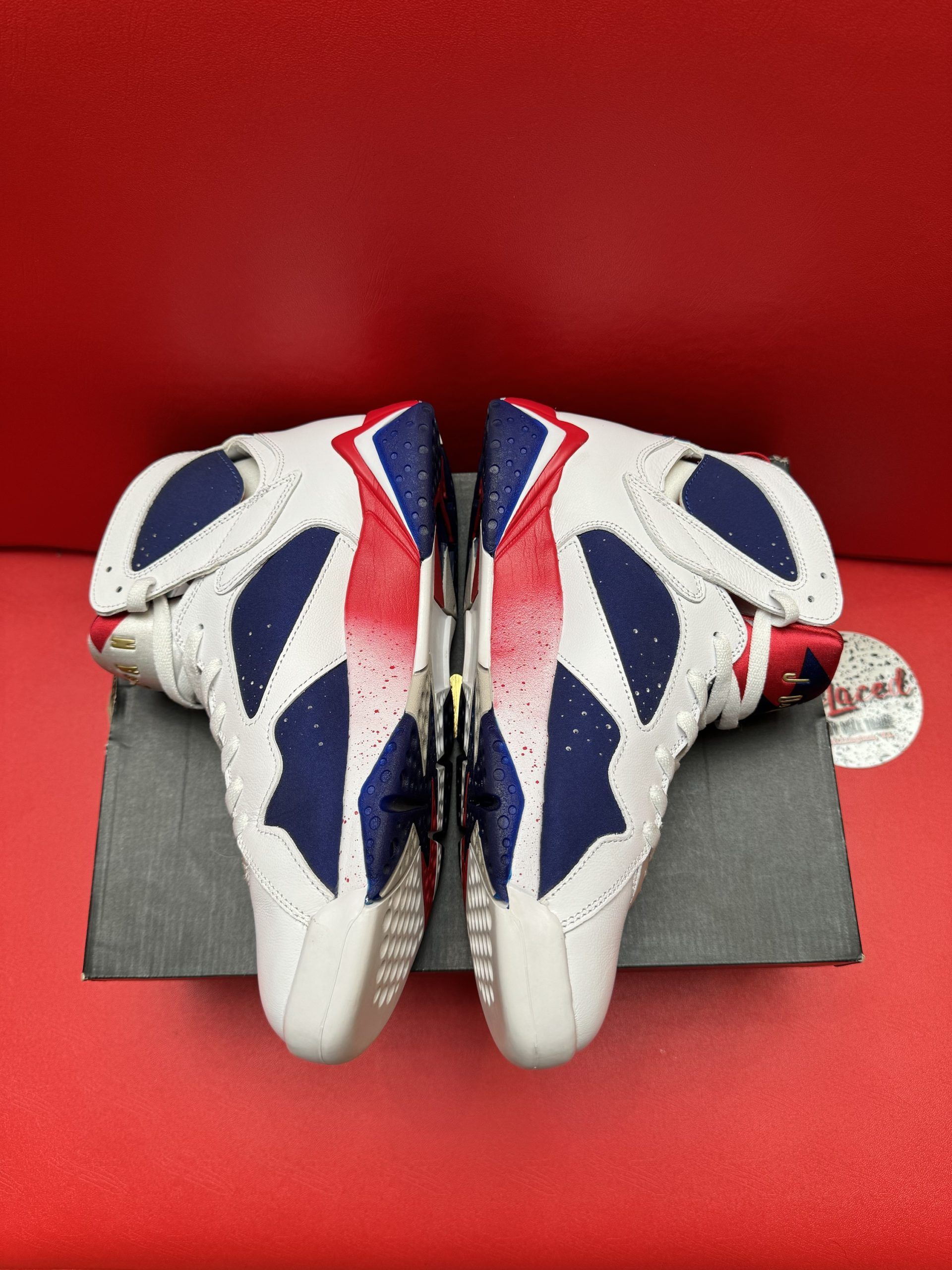 White, blue, and red Air Jordan 7 sneakers on a box, sole up, against a red background.