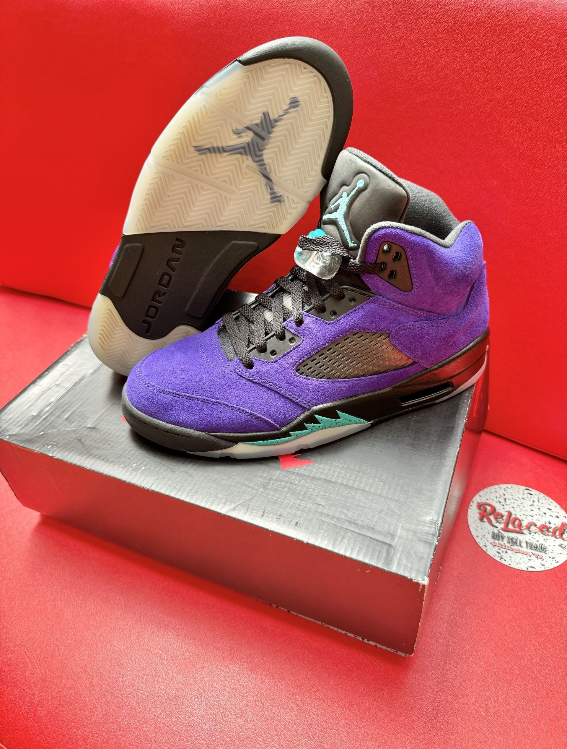 Purple and black Air Jordan 5 sneakers on a box, sole up, with a red background.