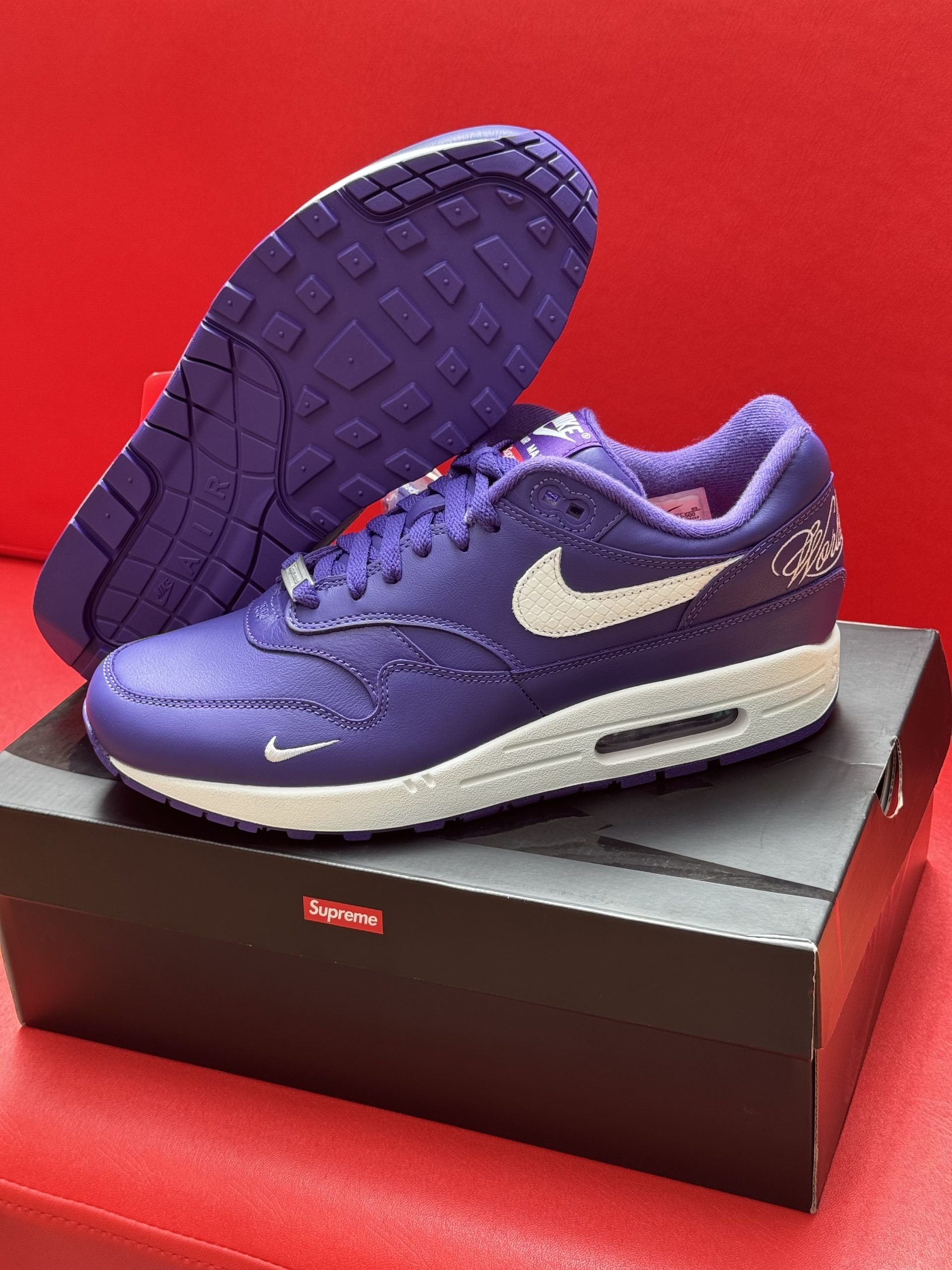 Purple Nike Air Max 1 sneakers with white accents and sole, on top of a Supreme box.