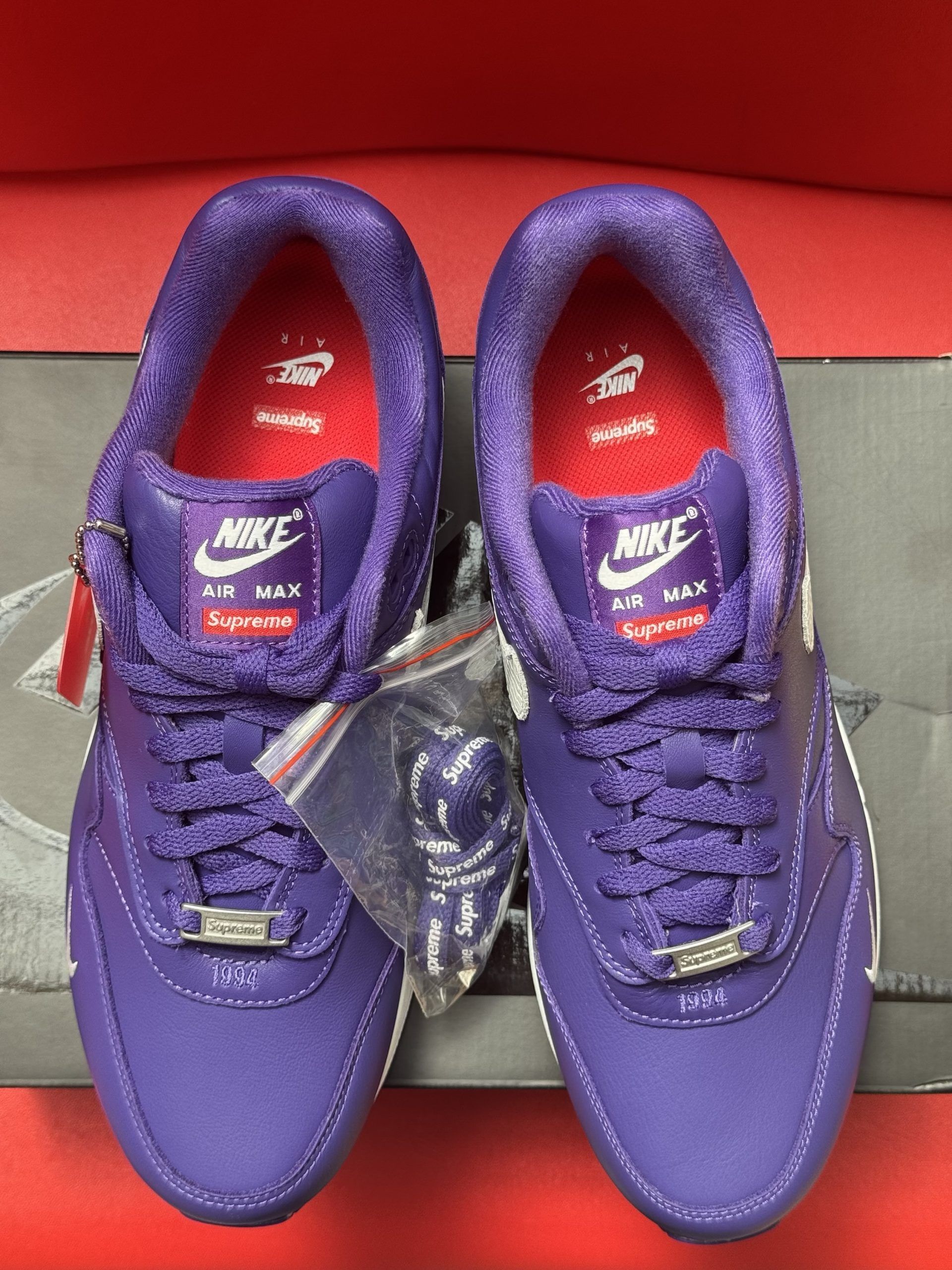 Purple Nike Air Max 1 sneakers with white accents and sole, on top of a Supreme box.