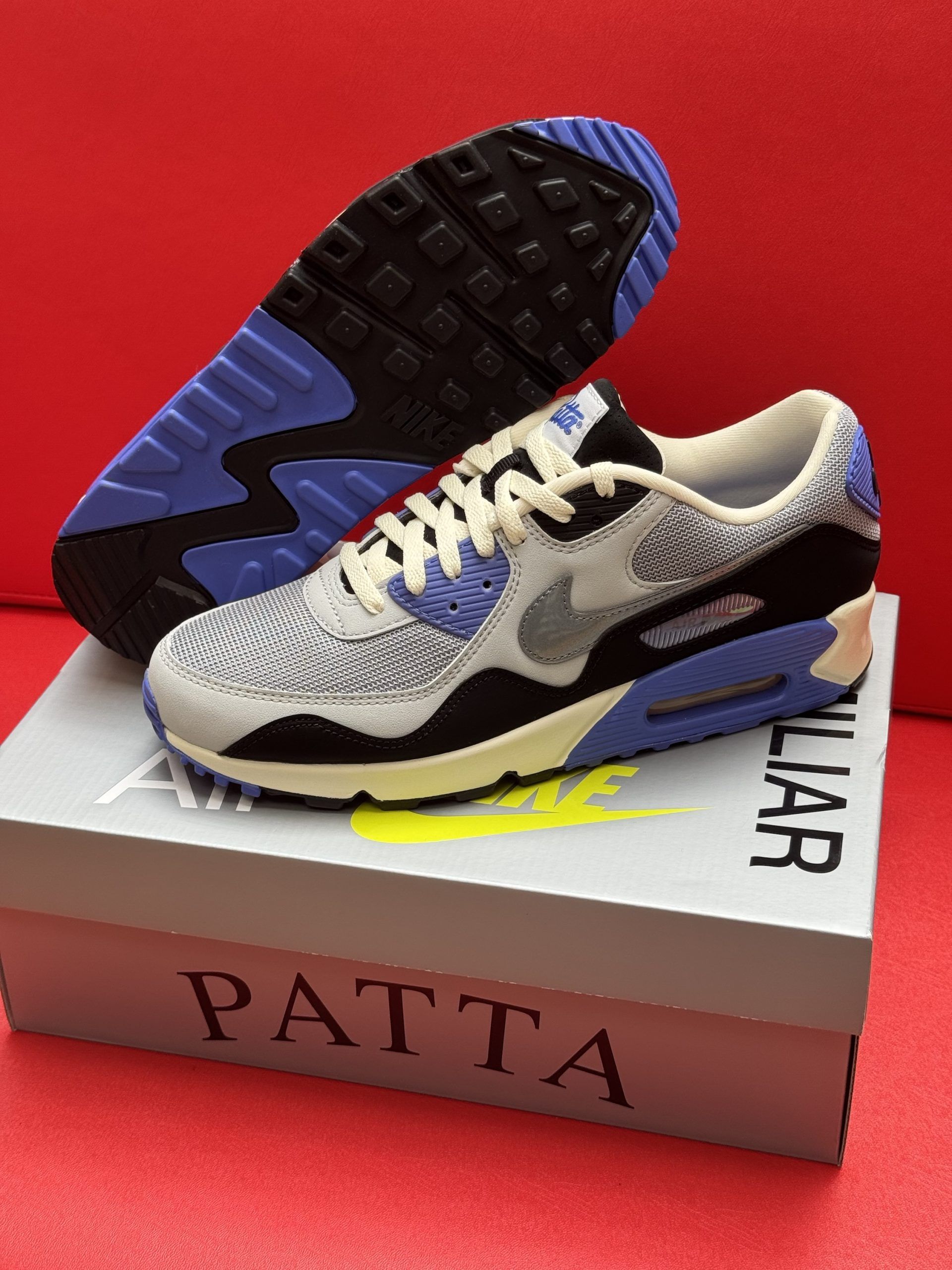Nike Air Max 90 sneakers, black and blue, on a Patta box, red background.
