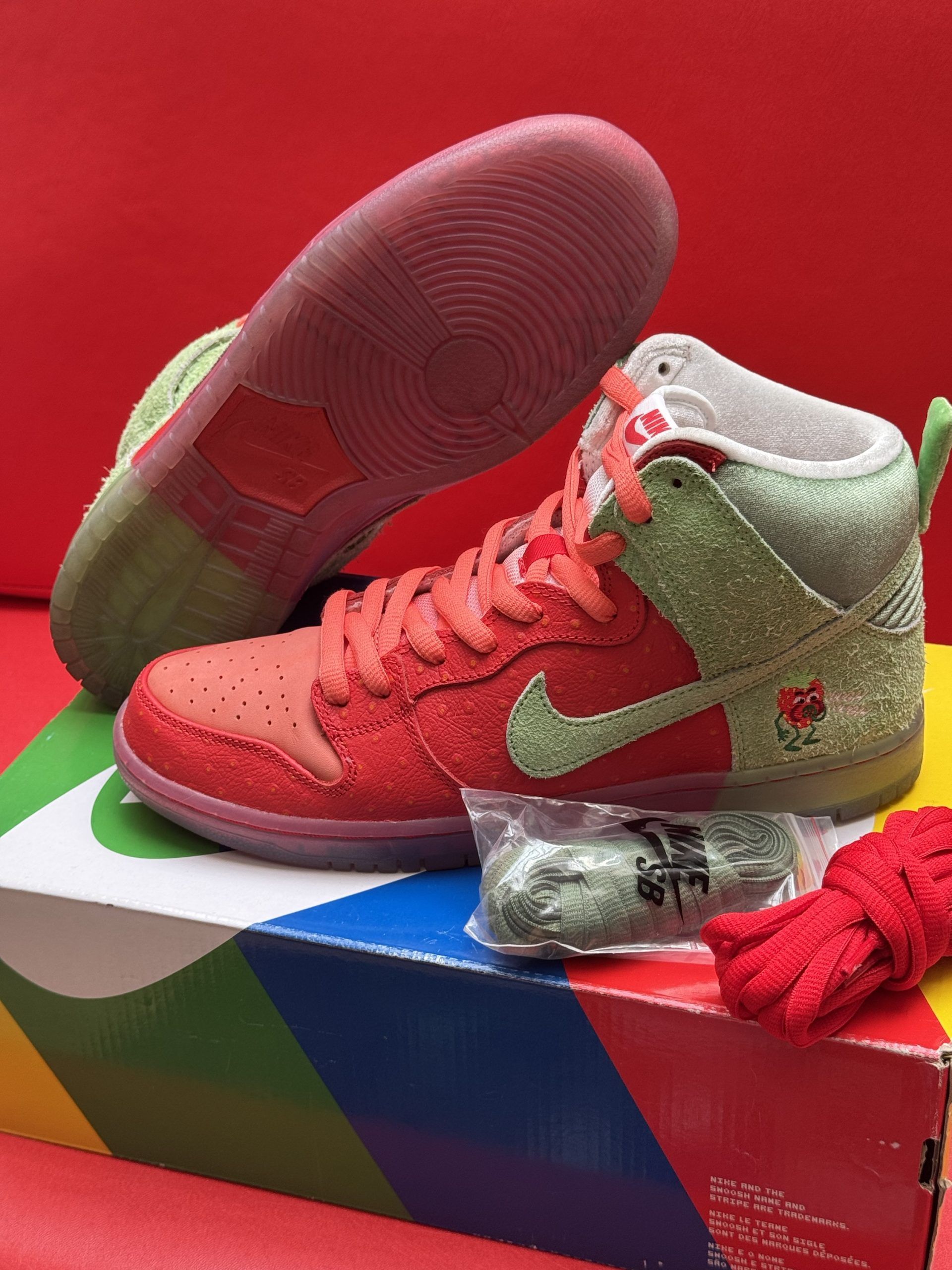 Red and green Nike Dunk High sneakers with extra laces and box on a red background.