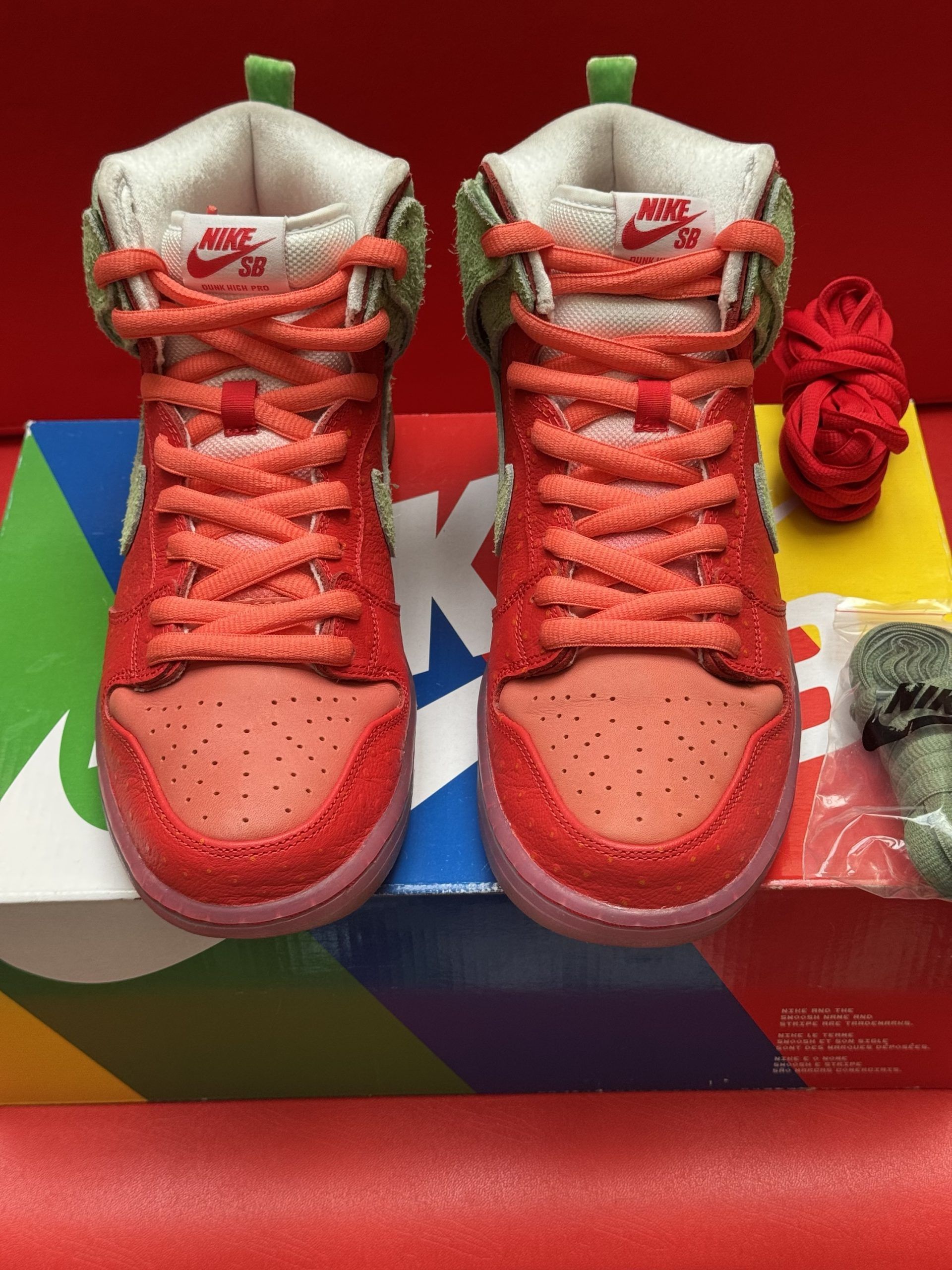 Red and green Nike Dunk High sneakers with extra laces and box on a red background.