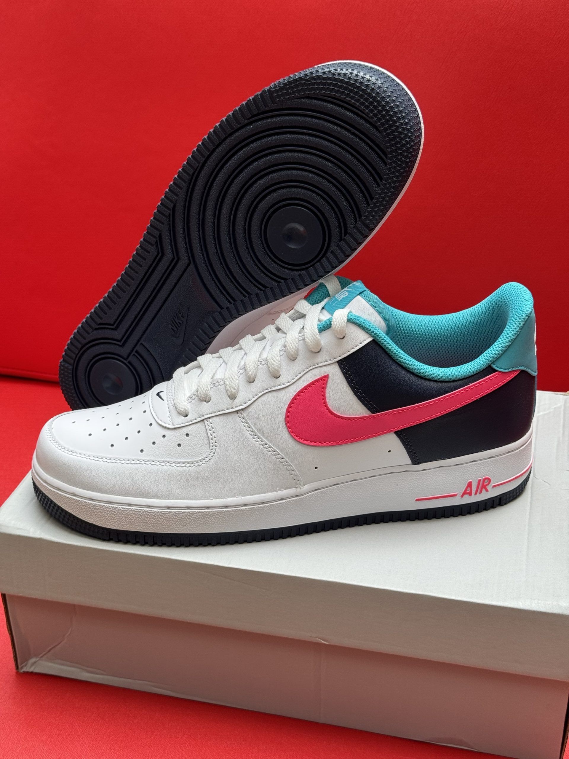 White Nike Air Force 1 sneaker with pink and teal accents, displayed on a shoebox.
