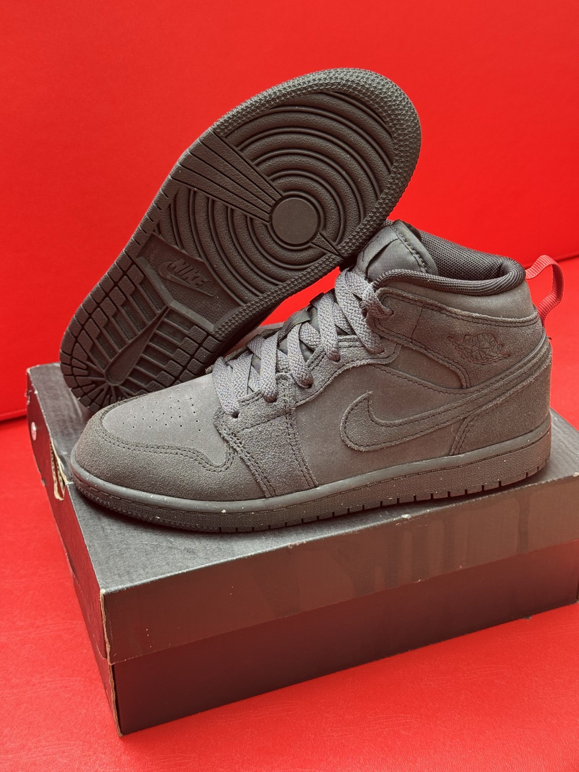 Black Jordan Mid sneakers on a box, sole exposed. Red background.