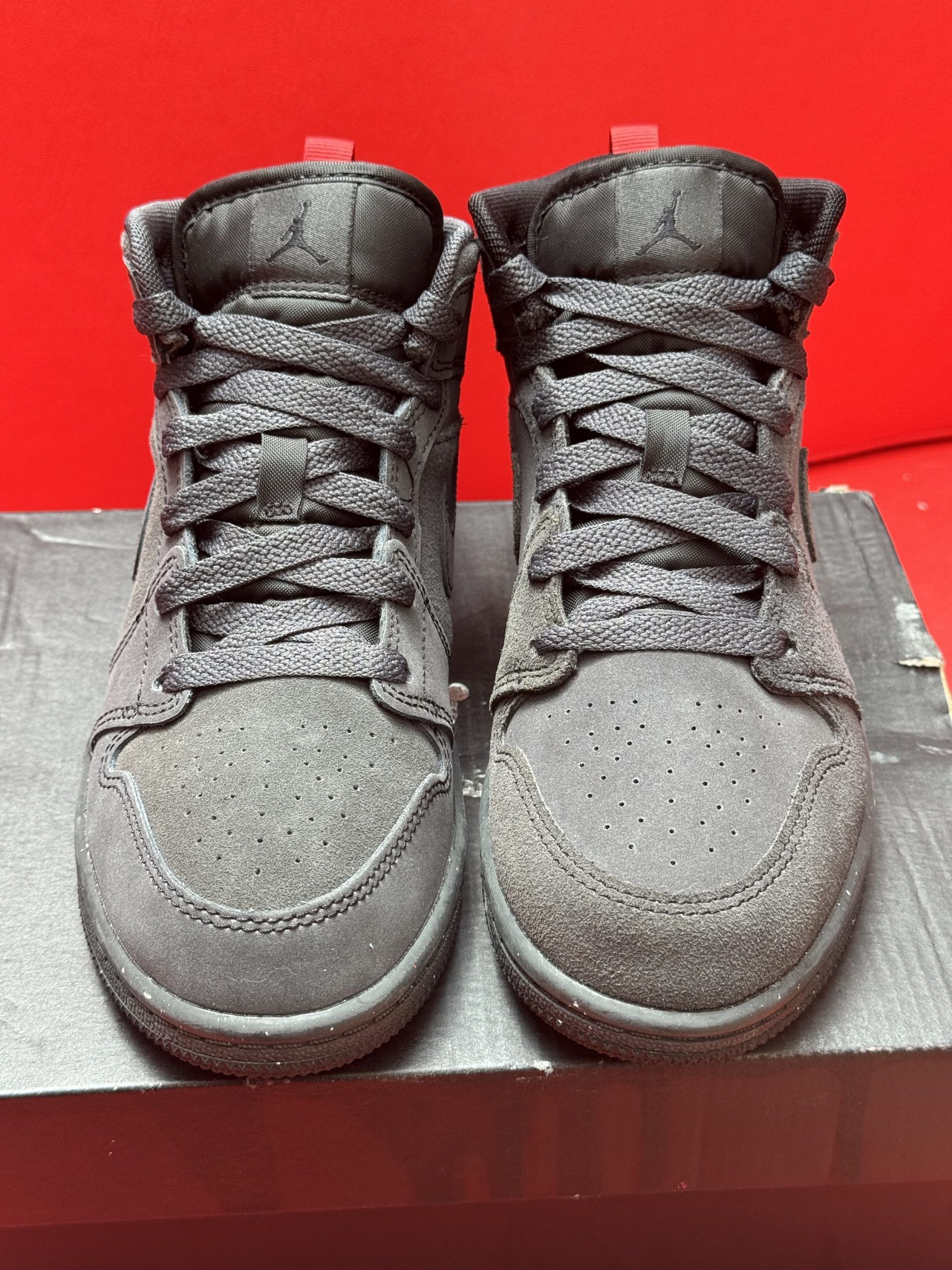 Black Jordan Mid sneakers on a box, sole exposed. Red background.