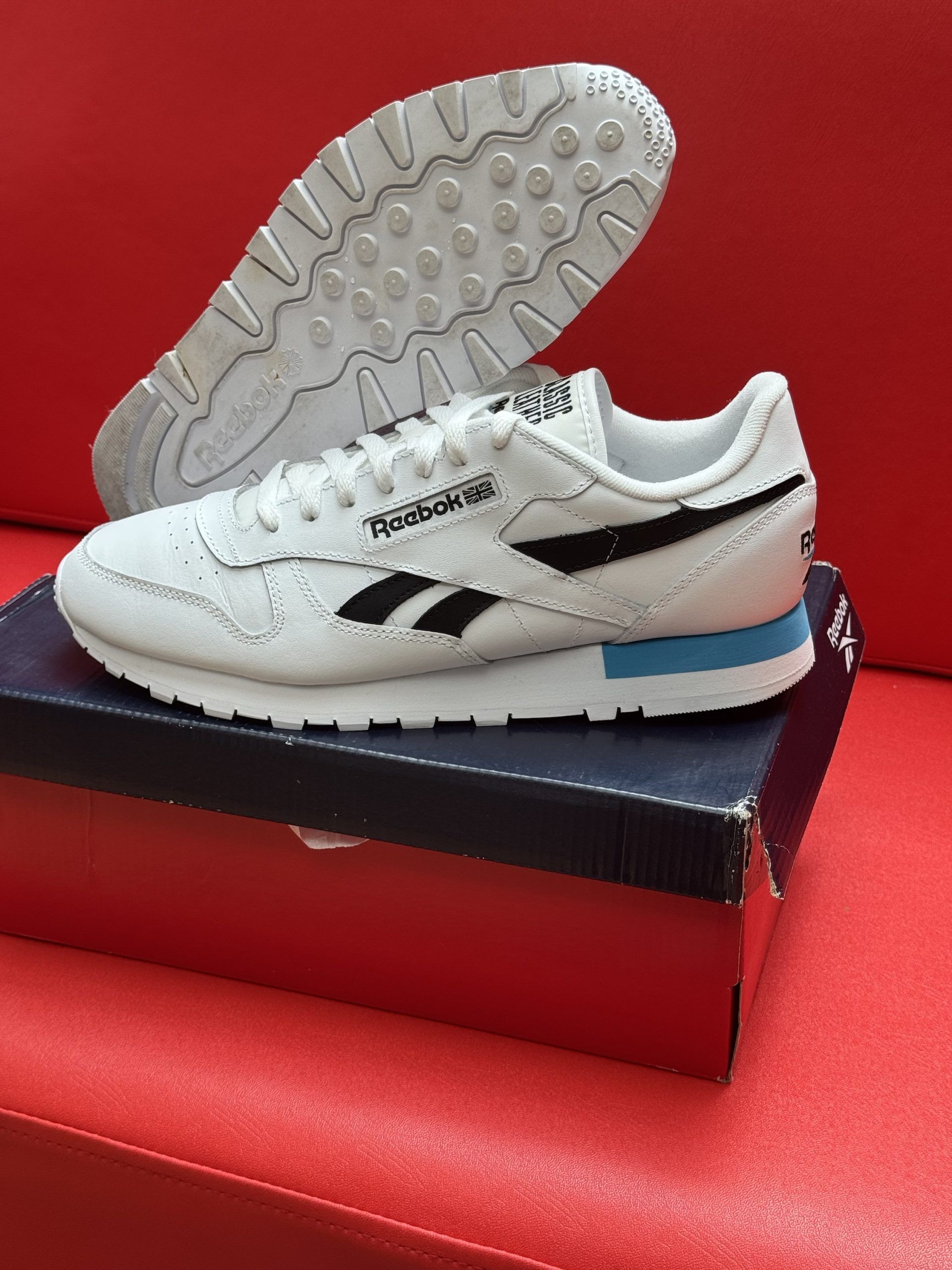 White Reebok sneakers with black stripes, sitting on a shoebox, with one sole visible on a red surface.