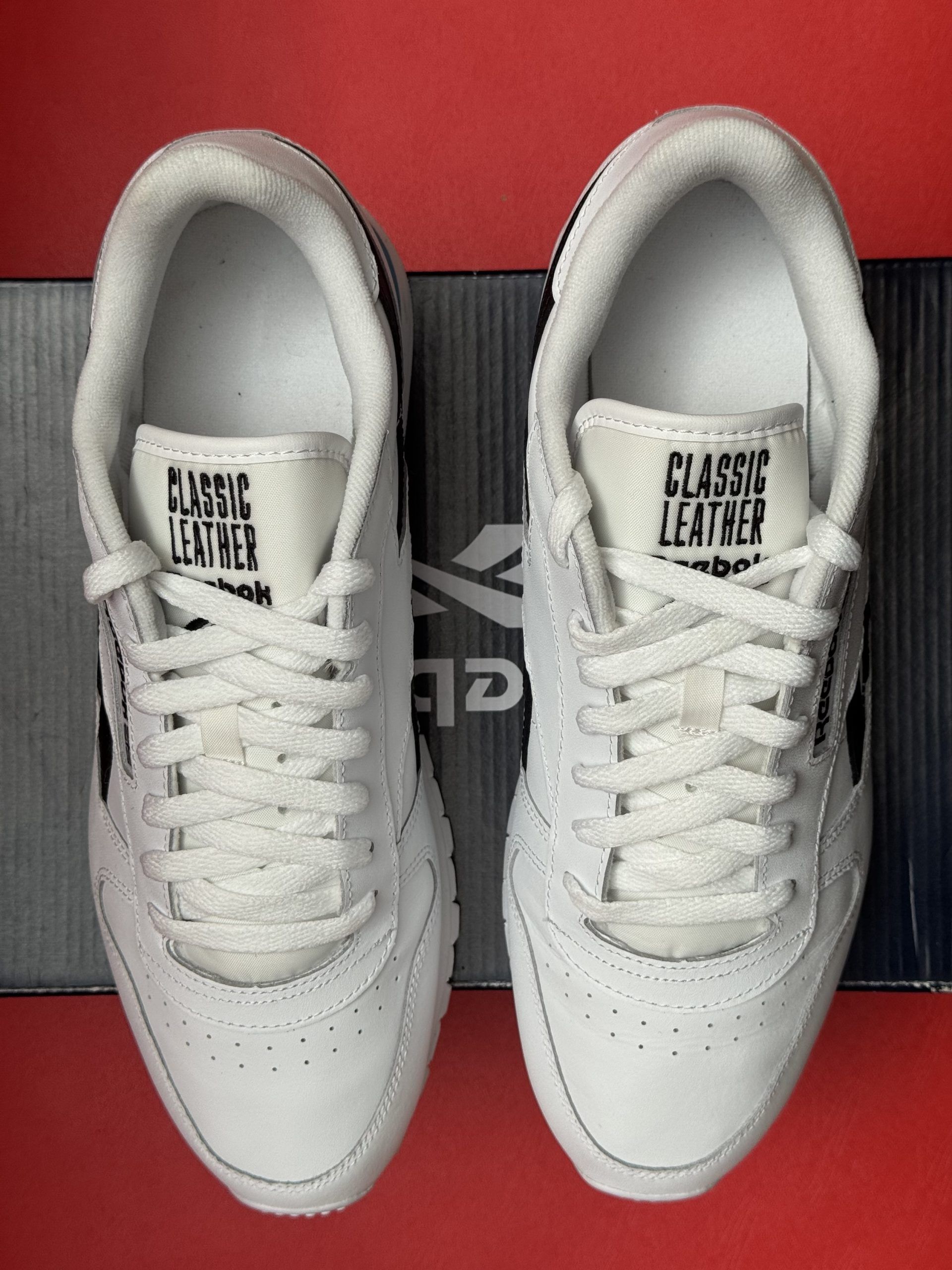 White Reebok sneakers with black stripes, sitting on a shoebox, with one sole visible on a red surface.