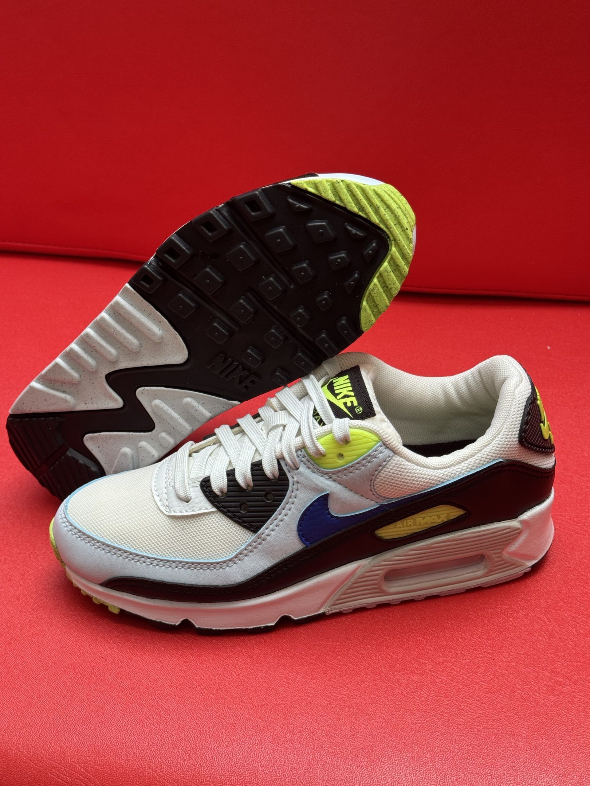 Nike Air Max 90 sneakers, white, black, blue, and yellow accents, one sole up, red background.