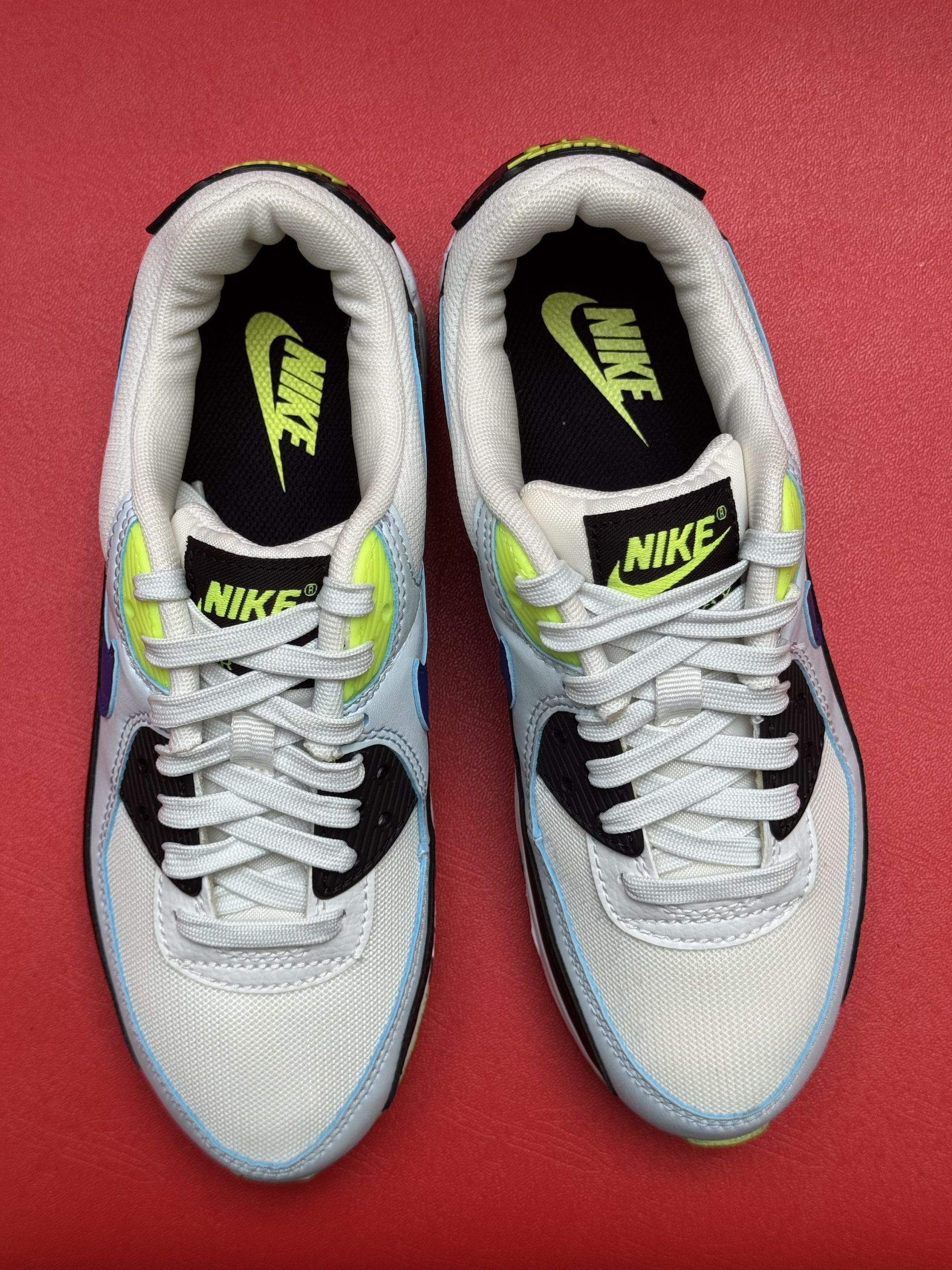 Nike Air Max 90 sneakers, white, black, blue, and yellow accents, one sole up, red background.