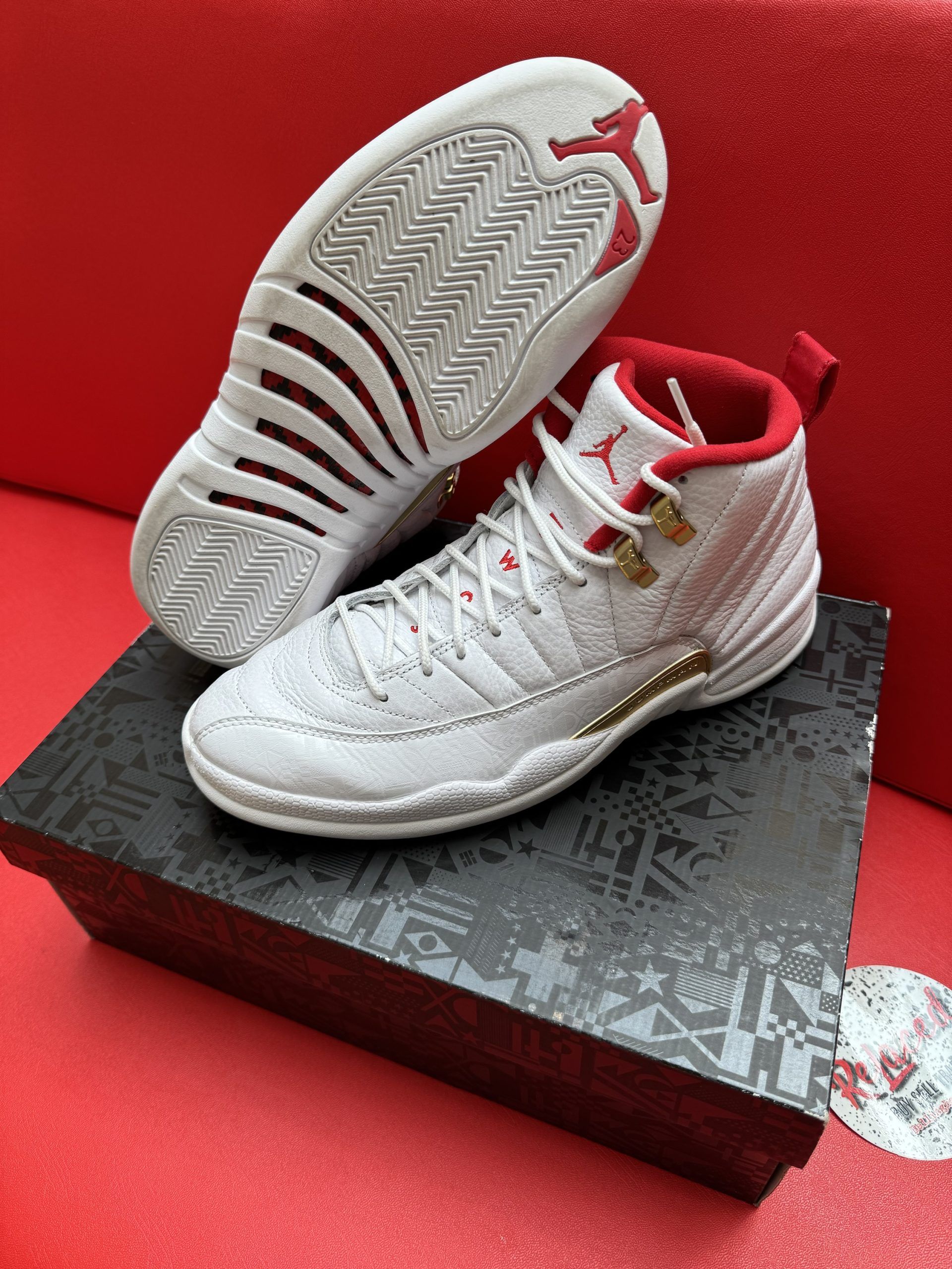 White and red Air Jordan 12 sneakers on a black patterned box, set against a red background.