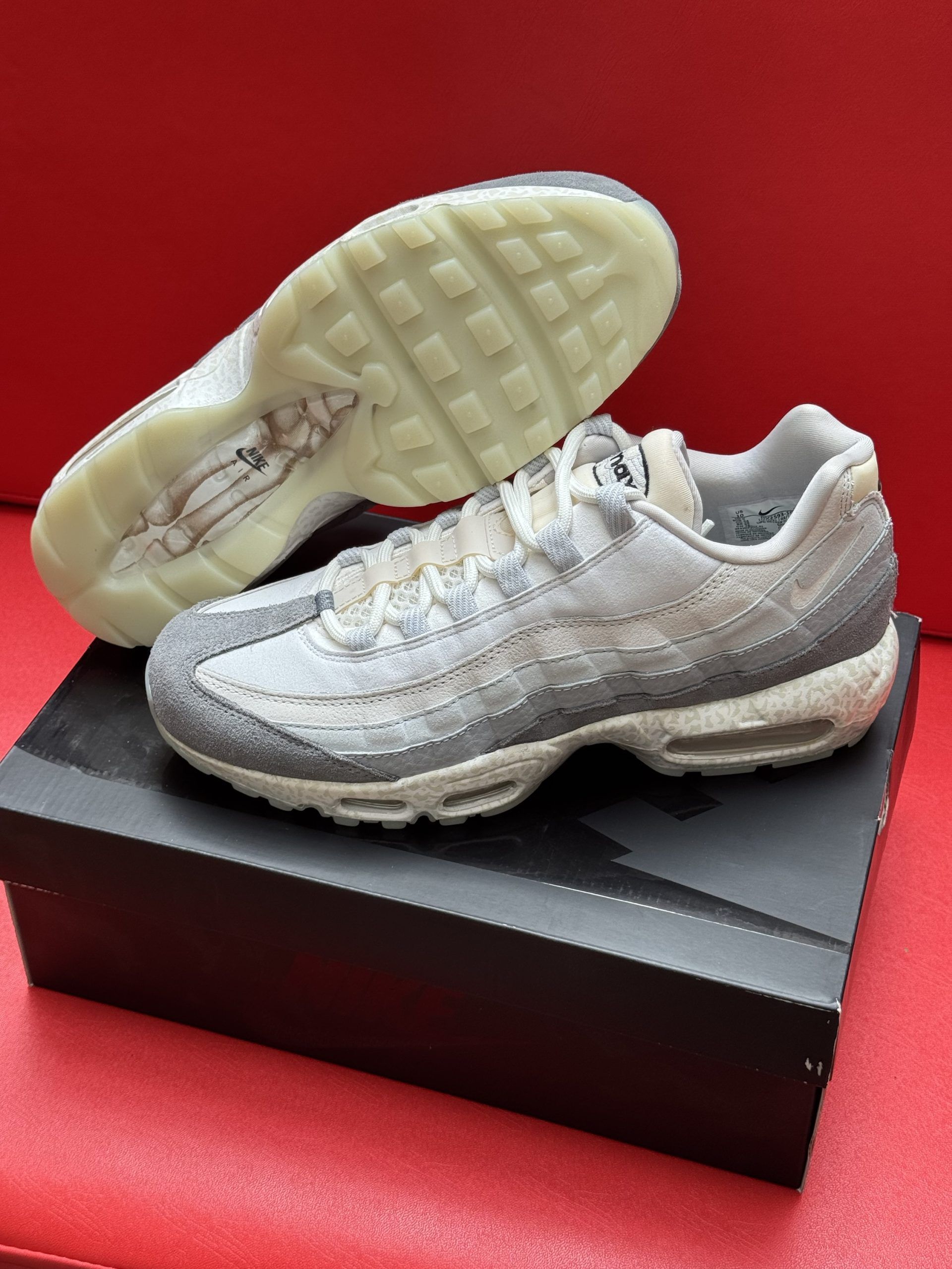Pair of gray and white Nike Air Max 95 sneakers on a black shoebox, against a red background.