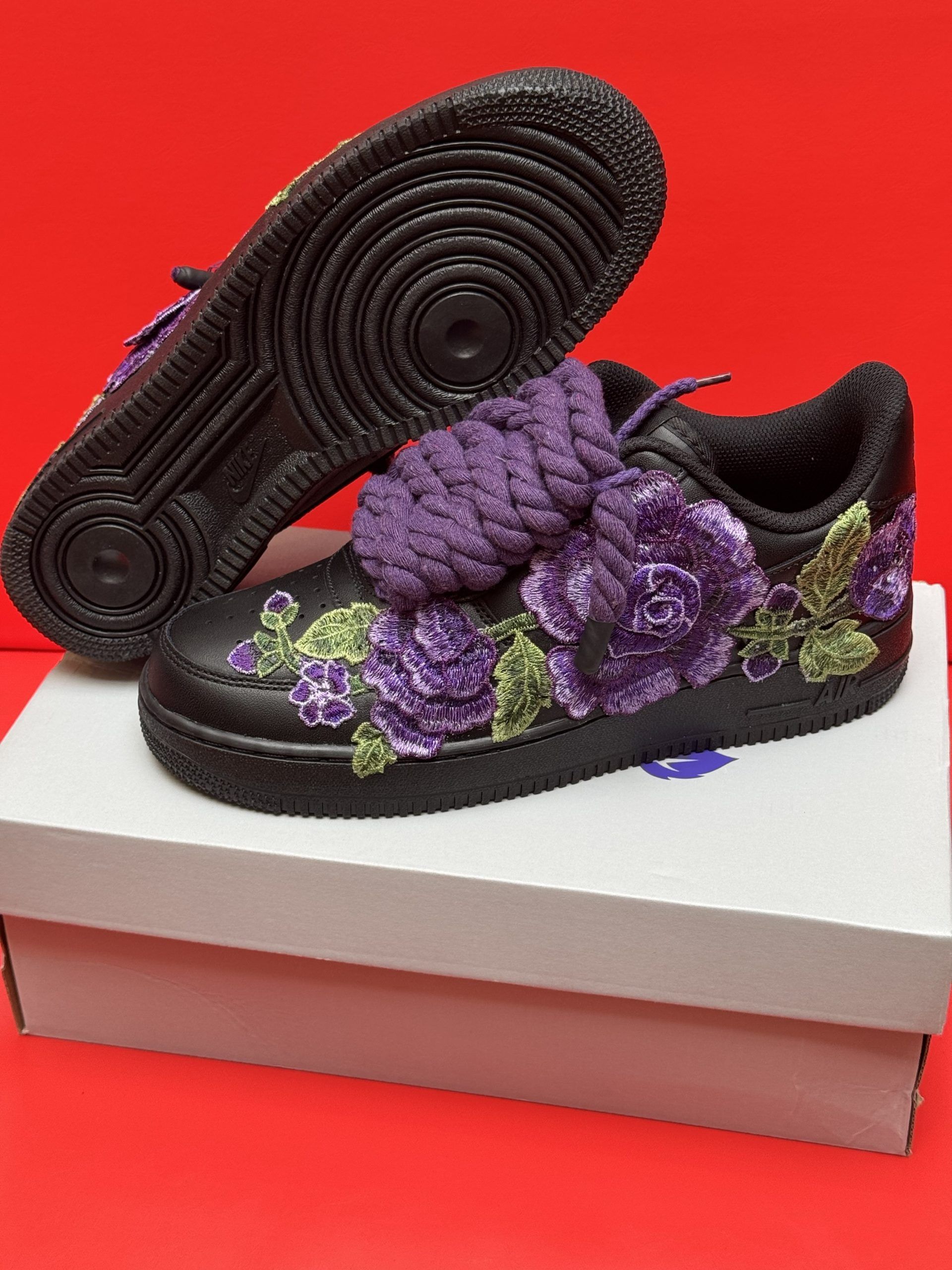 Black Air Force 1 sneakers with purple floral embroidery, a purple lace embellishment, and a black sole.