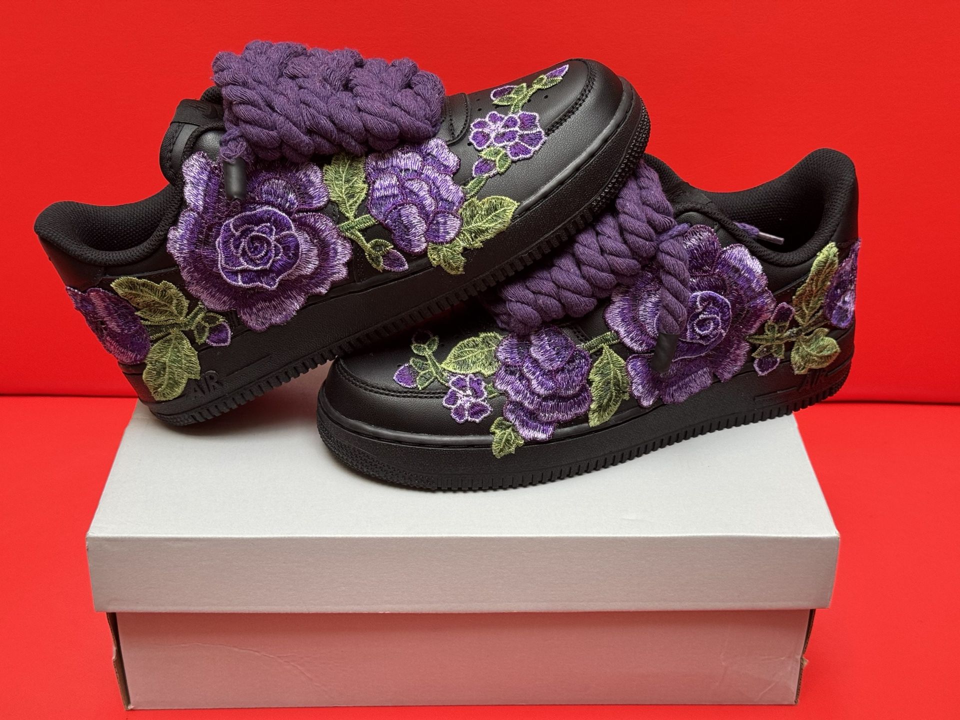 Black Air Force 1 sneakers with purple floral embroidery, a purple lace embellishment, and a black sole.