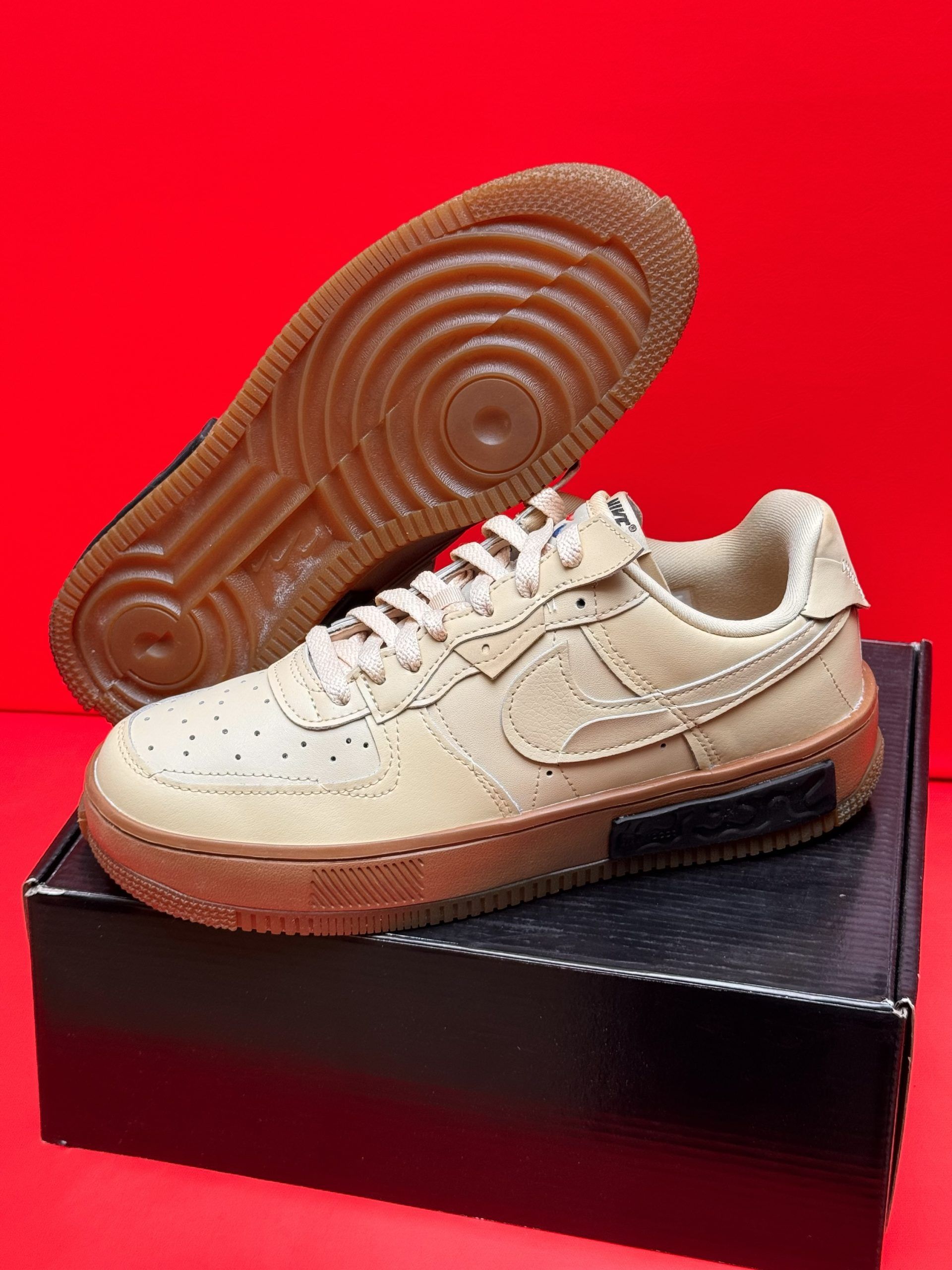 Beige Nike Air Force 1 sneakers with brown soles, sitting on a black box, against a red background.