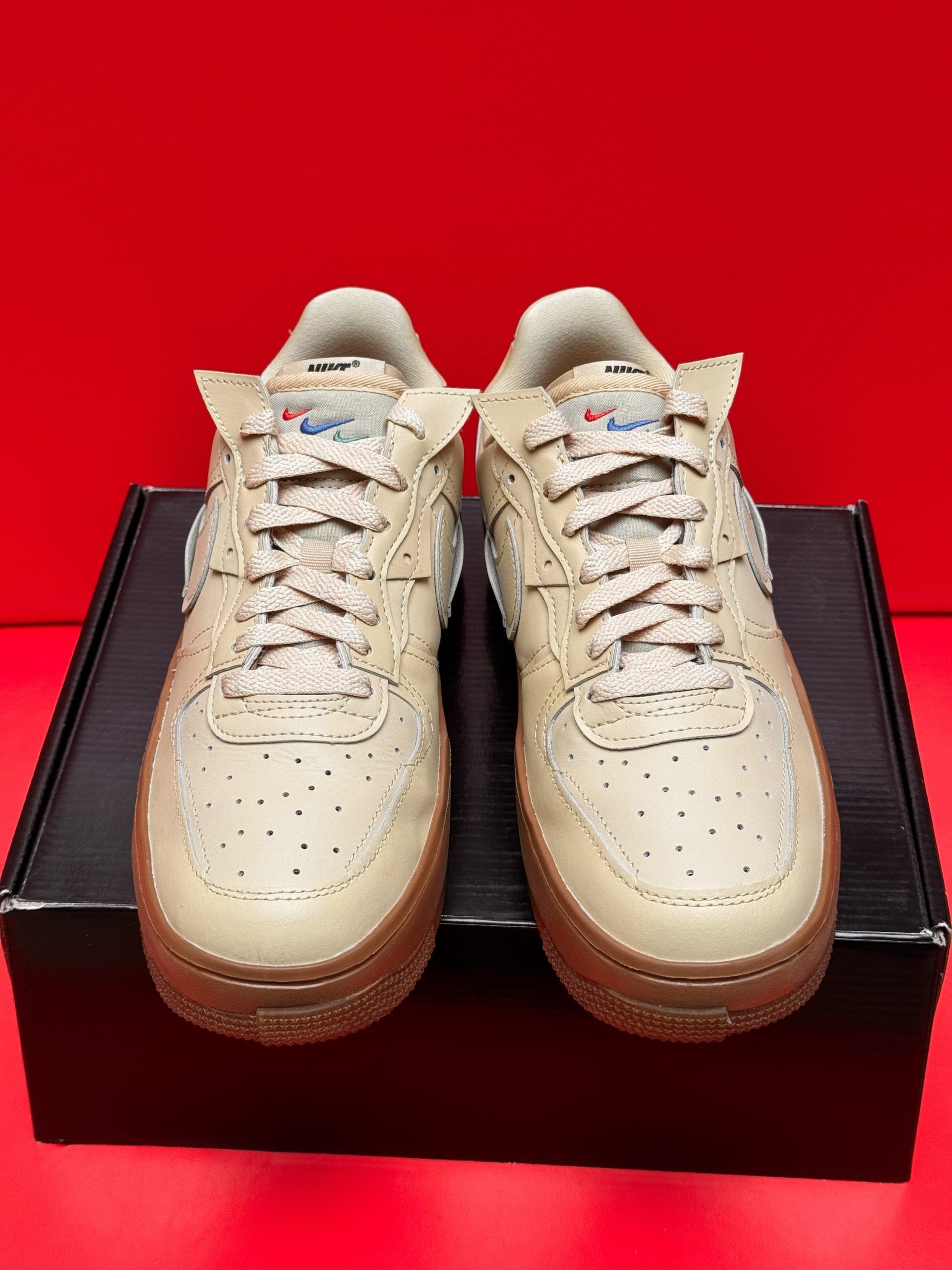 Beige Nike Air Force 1 sneakers with brown soles, sitting on a black box, against a red background.