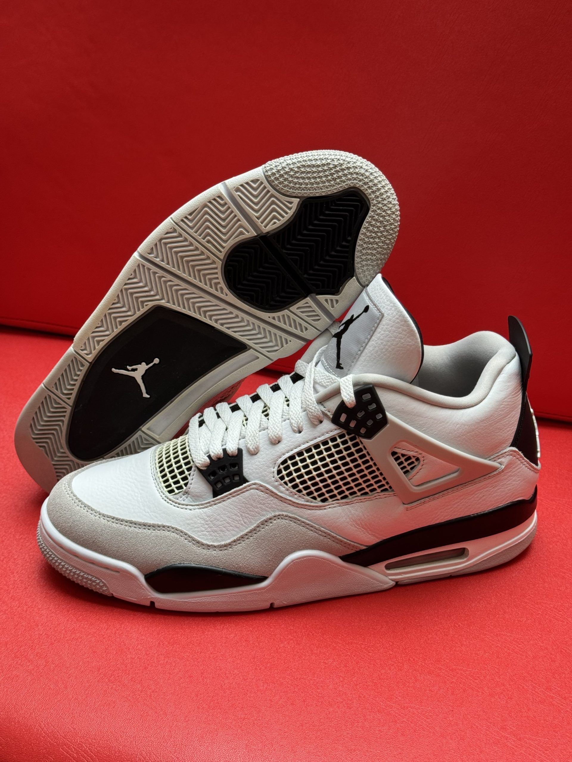White and gray Air Jordan 4 sneakers on a red surface, with one shoe turned to show the sole.