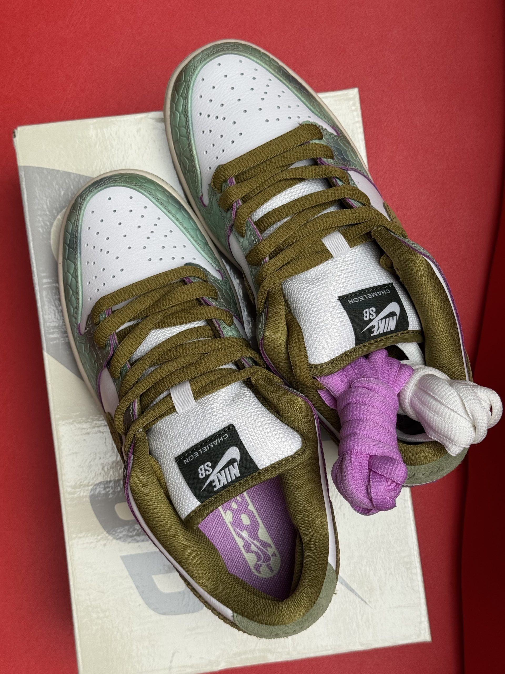 Nike SB Dunk Low sneakers in white, green, and brown, sitting on a shoebox against a red backdrop.
