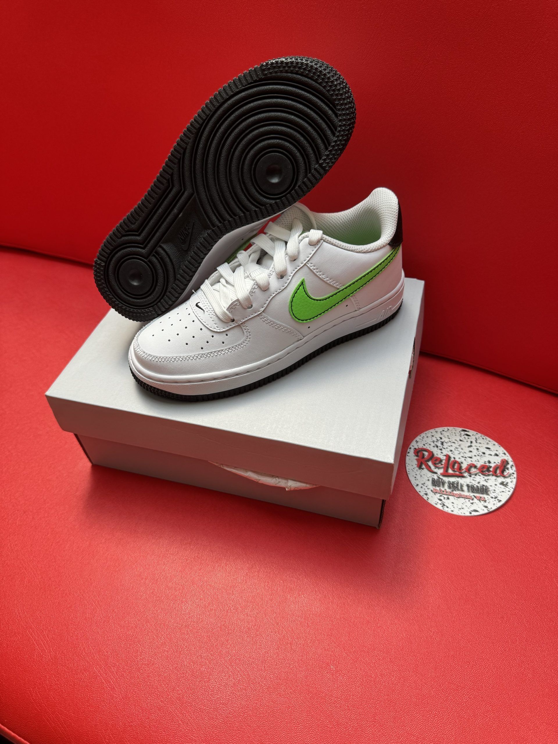 White Nike Air Force 1 sneakers with a green swoosh, resting on a shoebox. A black outsole is in the background on a red surface.