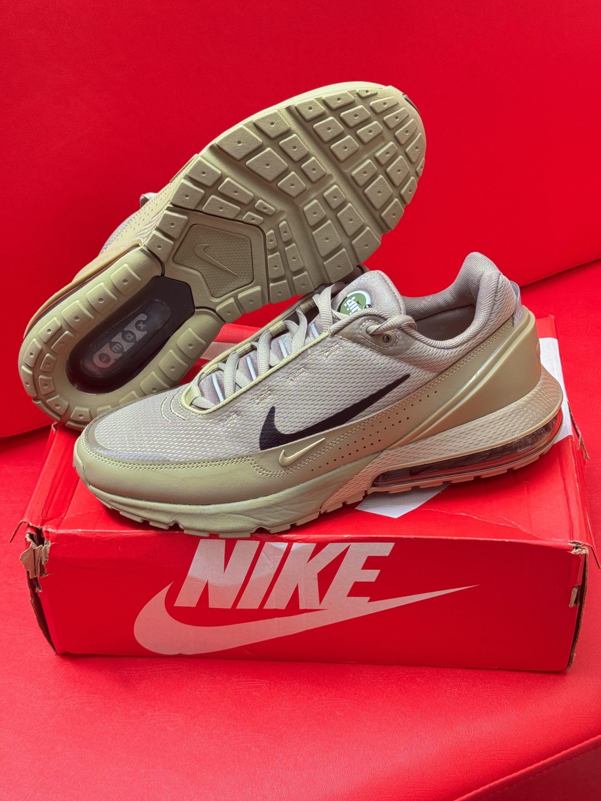 Nike Air Max shoes on a red Nike box, against a red background. The shoes are tan with a black swoosh.