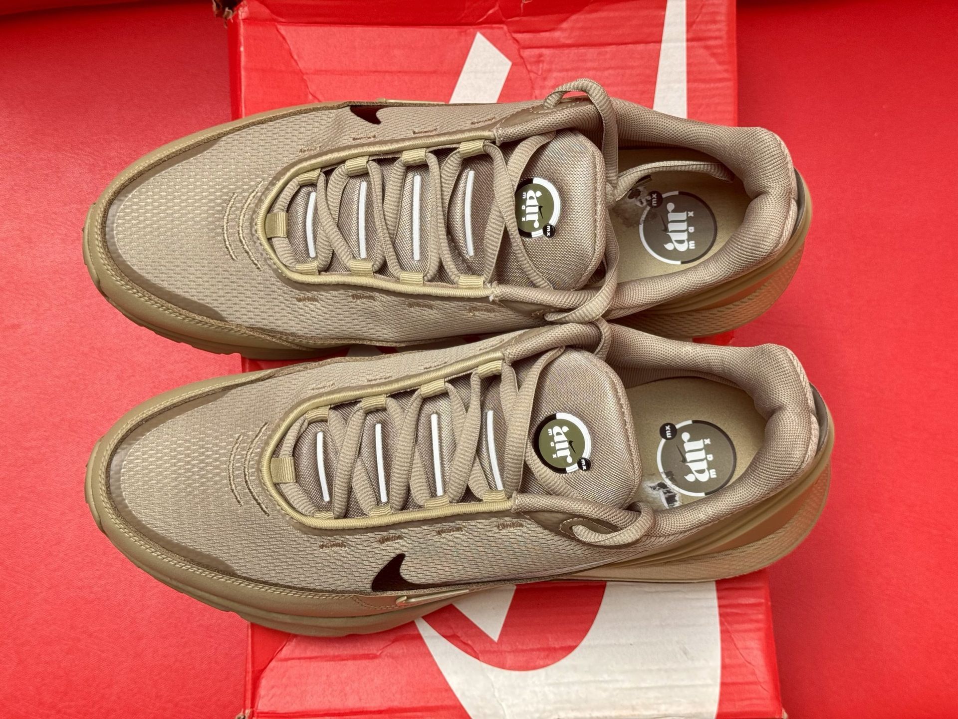 Nike Air Max shoes on a red Nike box, against a red background. The shoes are tan with a black swoosh.