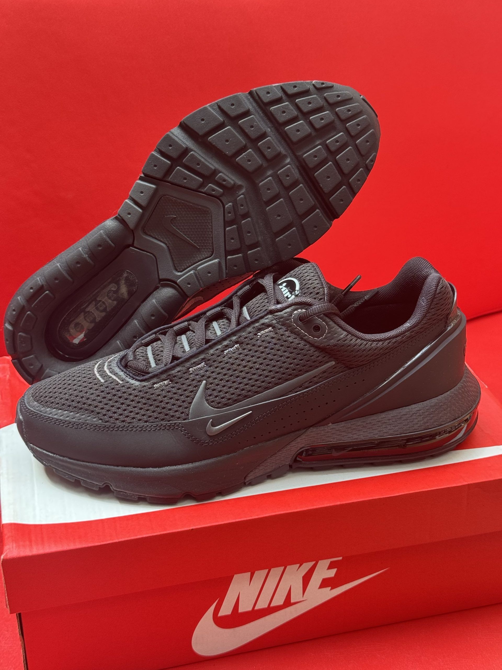 Black Nike sneakers on a red Nike shoebox, another shoe sole up in the background, all against a red backdrop.