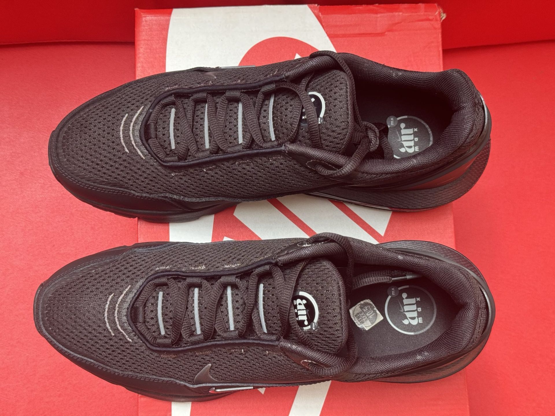 Black Nike sneakers on a red Nike shoebox, another shoe sole up in the background, all against a red backdrop.