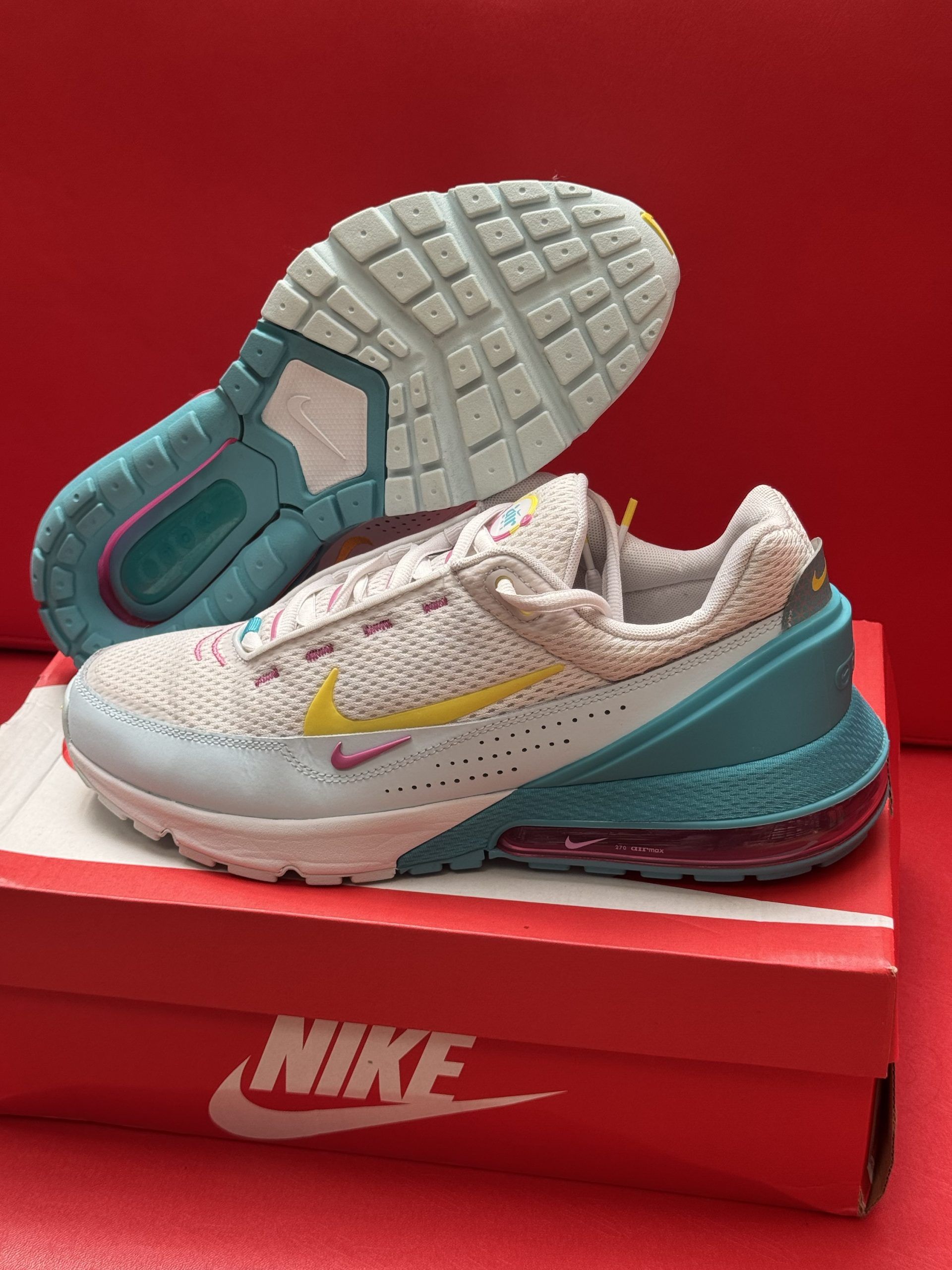 White and blue Nike Air Max shoes with yellow and pink accents, set on a Nike box against a red backdrop.