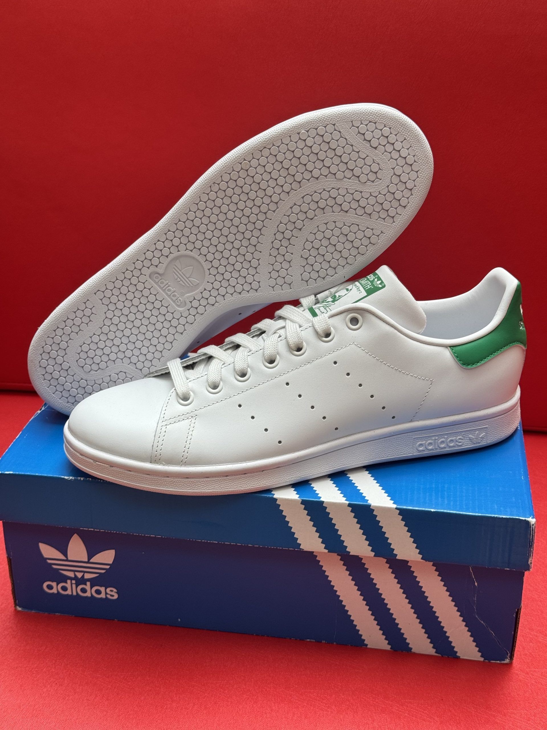 White Adidas Stan Smith sneakers with green heel on a blue Adidas box, one shoe upside down.