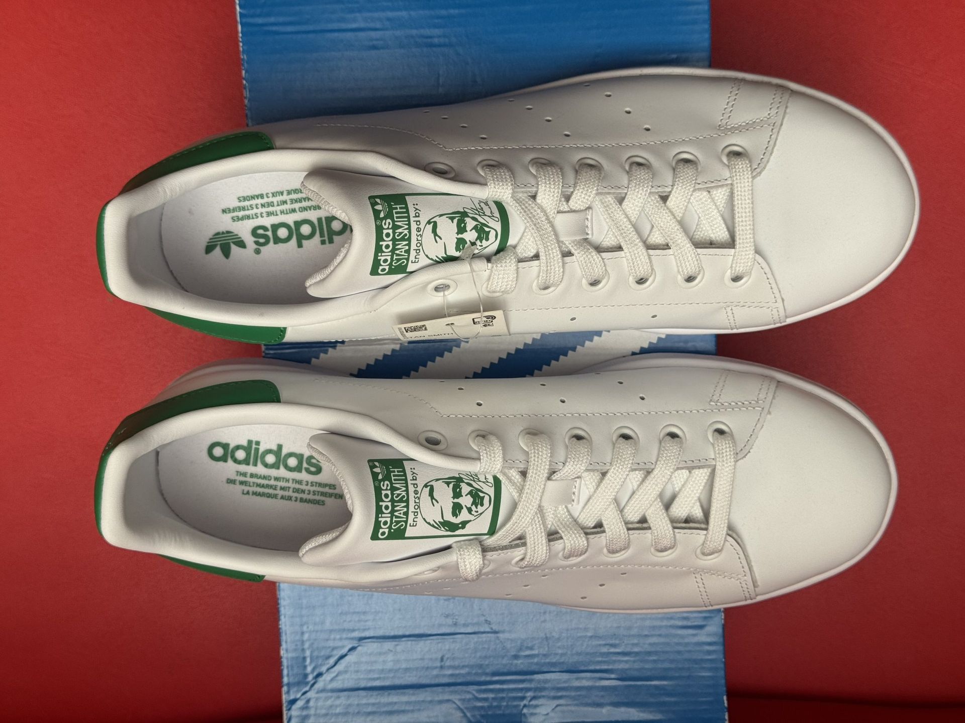 White Adidas Stan Smith sneakers with green heel on a blue Adidas box, one shoe upside down.