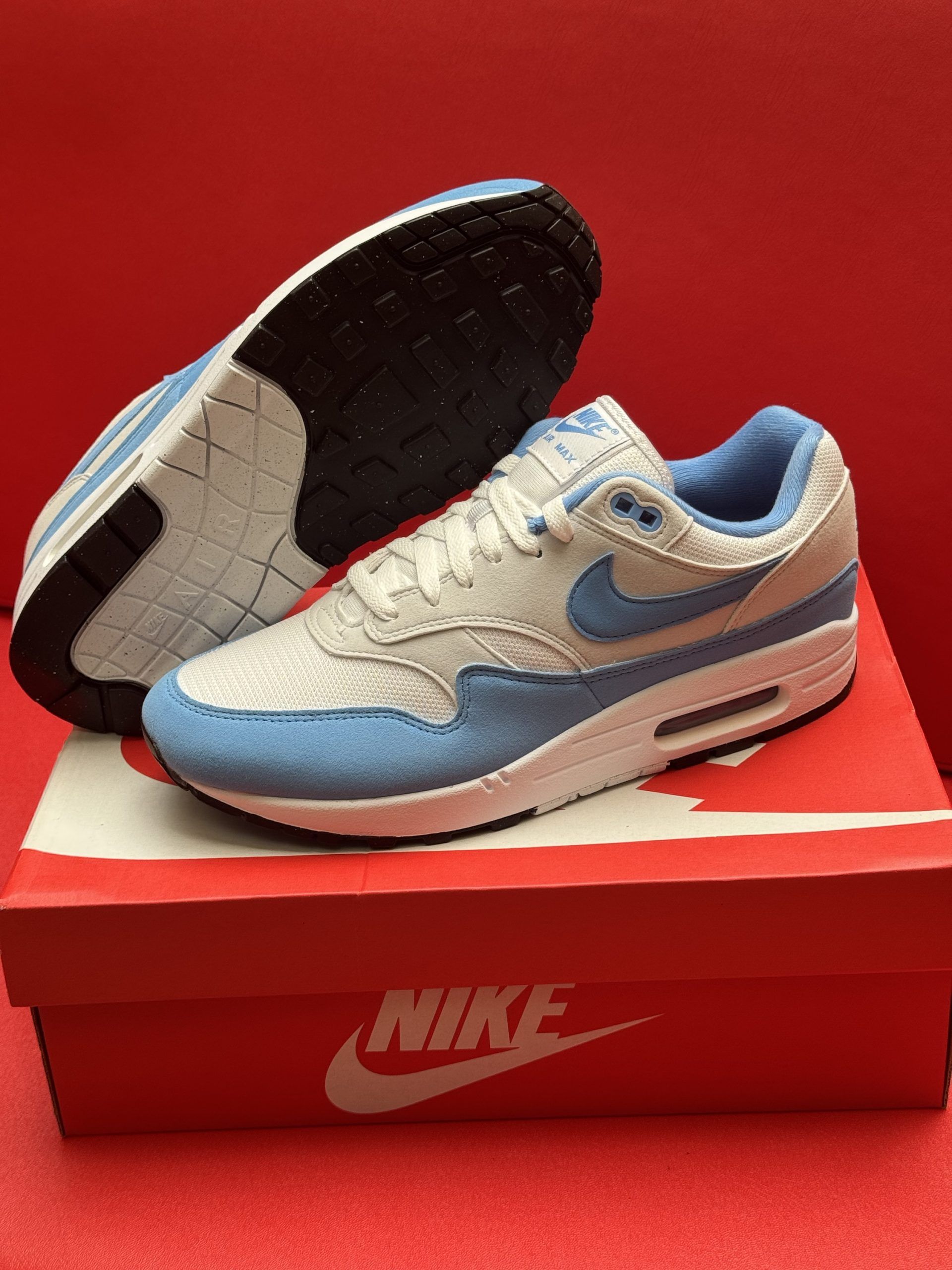 Light blue and white Nike Air Max 1 sneakers on a red Nike shoebox, with a red background.