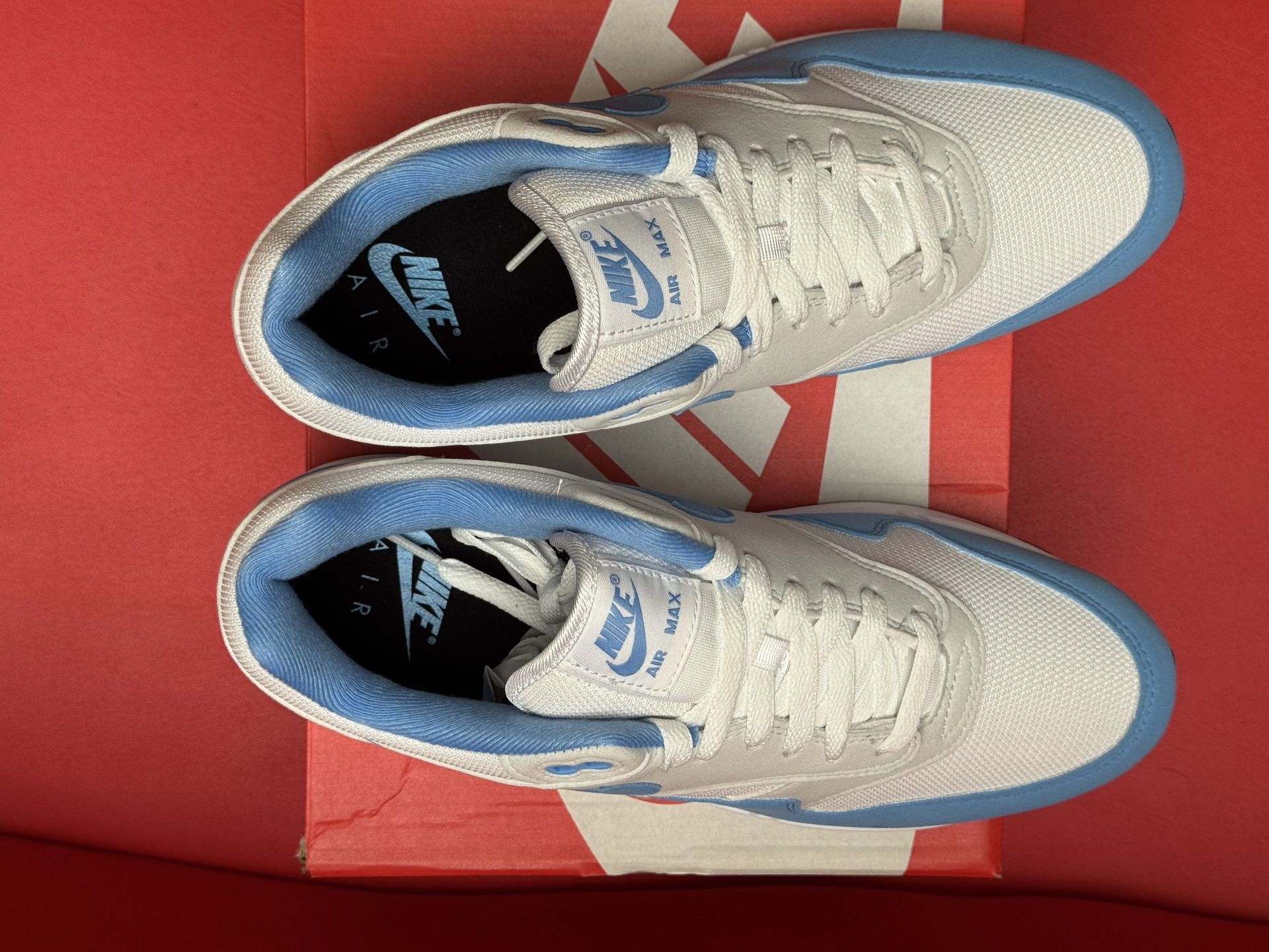 Light blue and white Nike Air Max 1 sneakers on a red Nike shoebox, with a red background.