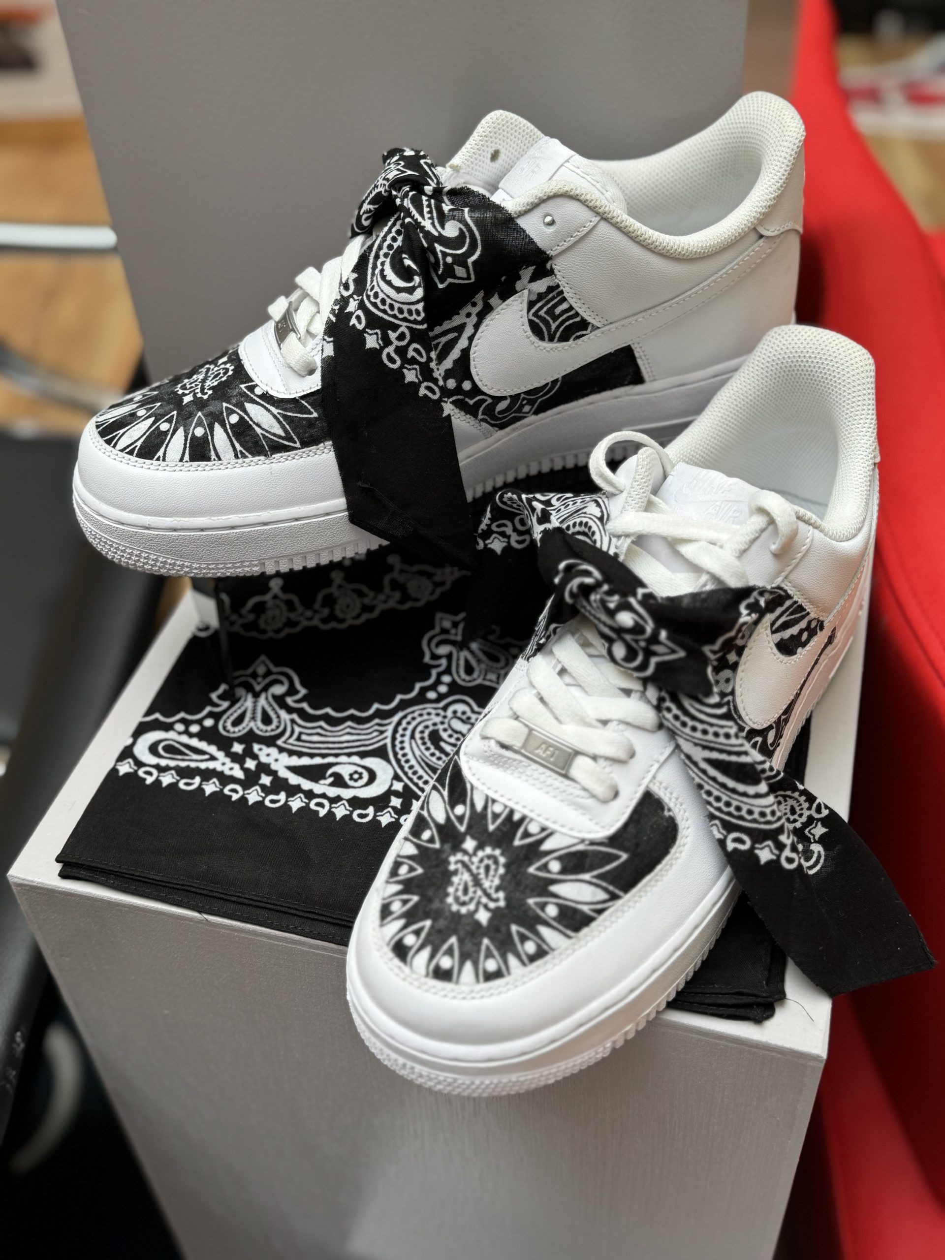 White Nike Air Force 1 sneakers customized with black bandana accents, sitting on a white pedestal with a matching bandana.