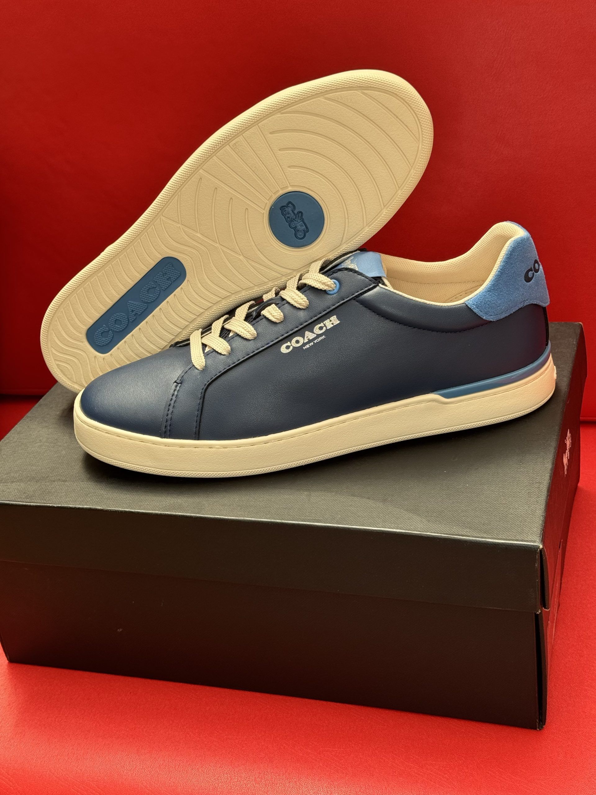 Blue Coach sneakers on a black box, with the sole visible; set against a red background.