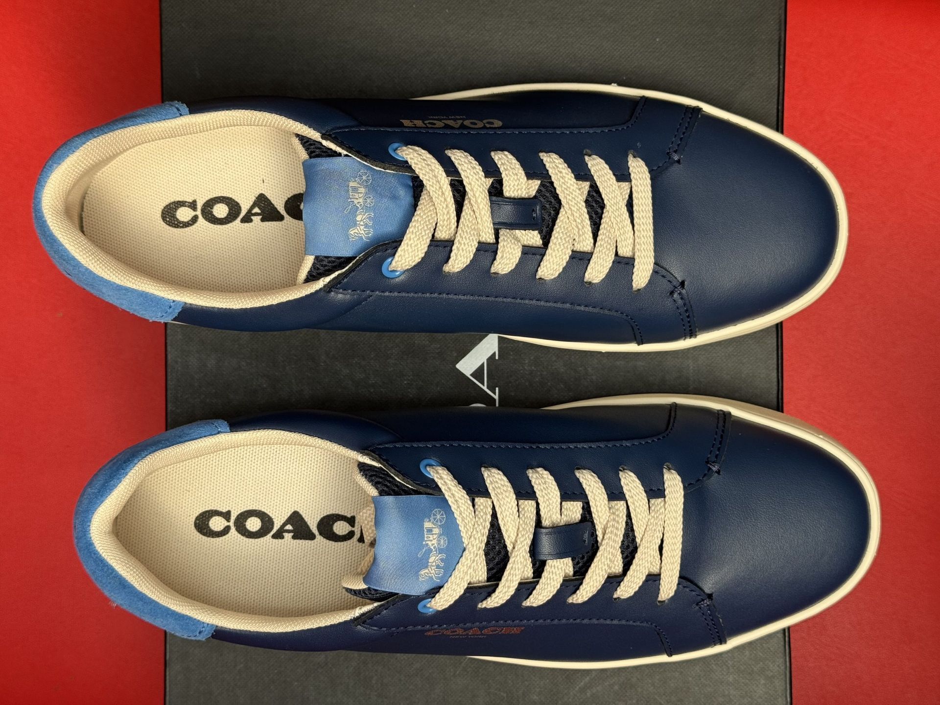 Blue Coach sneakers on a black box, with the sole visible; set against a red background.