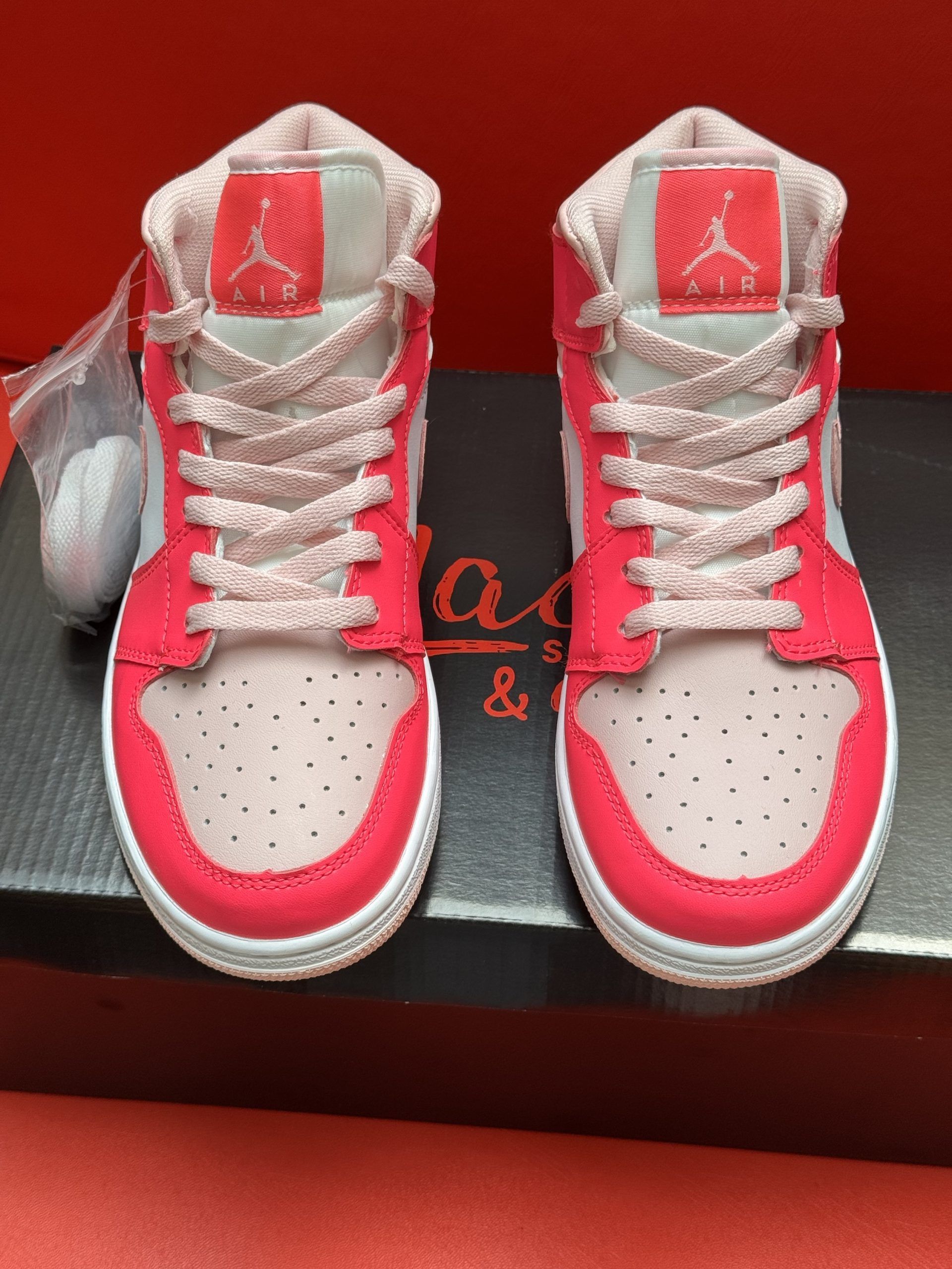 Pink and white Air Jordan sneakers on a black box, with a red background. The sole is visible.