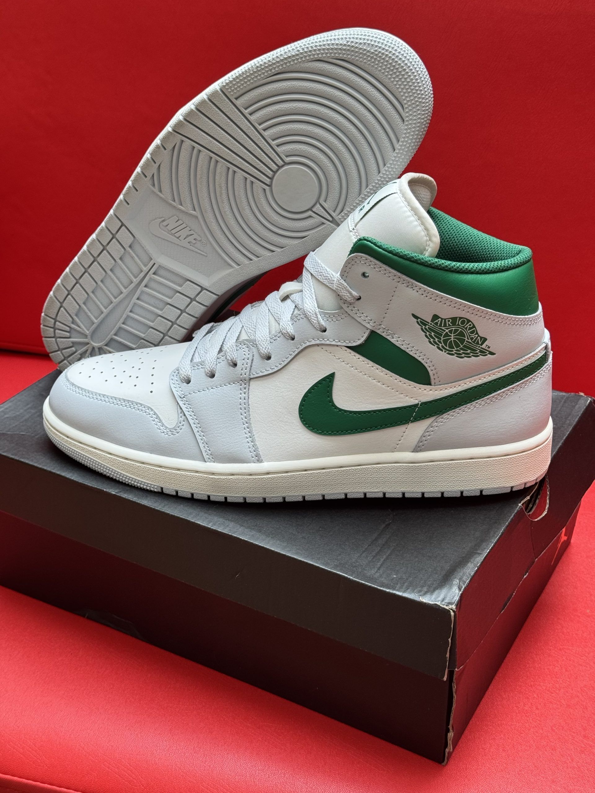 White and green Air Jordan 1 sneaker on black box, red background. Sole up.