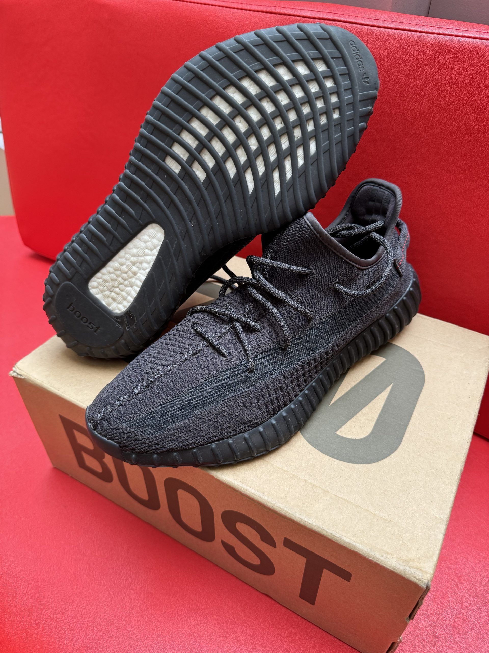 Pair of black Yeezy Boost 350 V2 sneakers with their box, set on a red surface. One shoe is inverted, showing its sole.