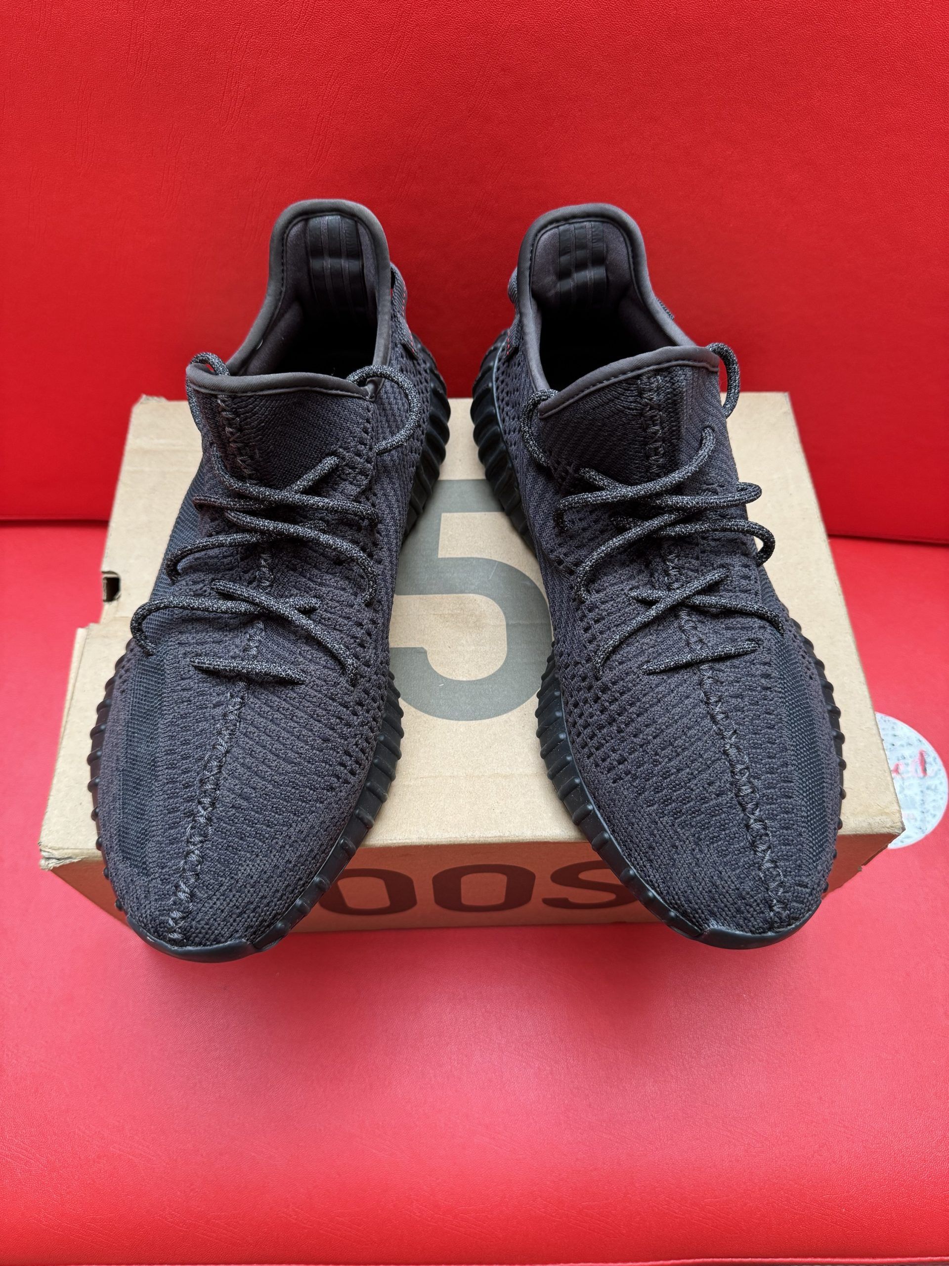 Pair of black Yeezy Boost 350 V2 sneakers with their box, set on a red surface. One shoe is inverted, showing its sole.