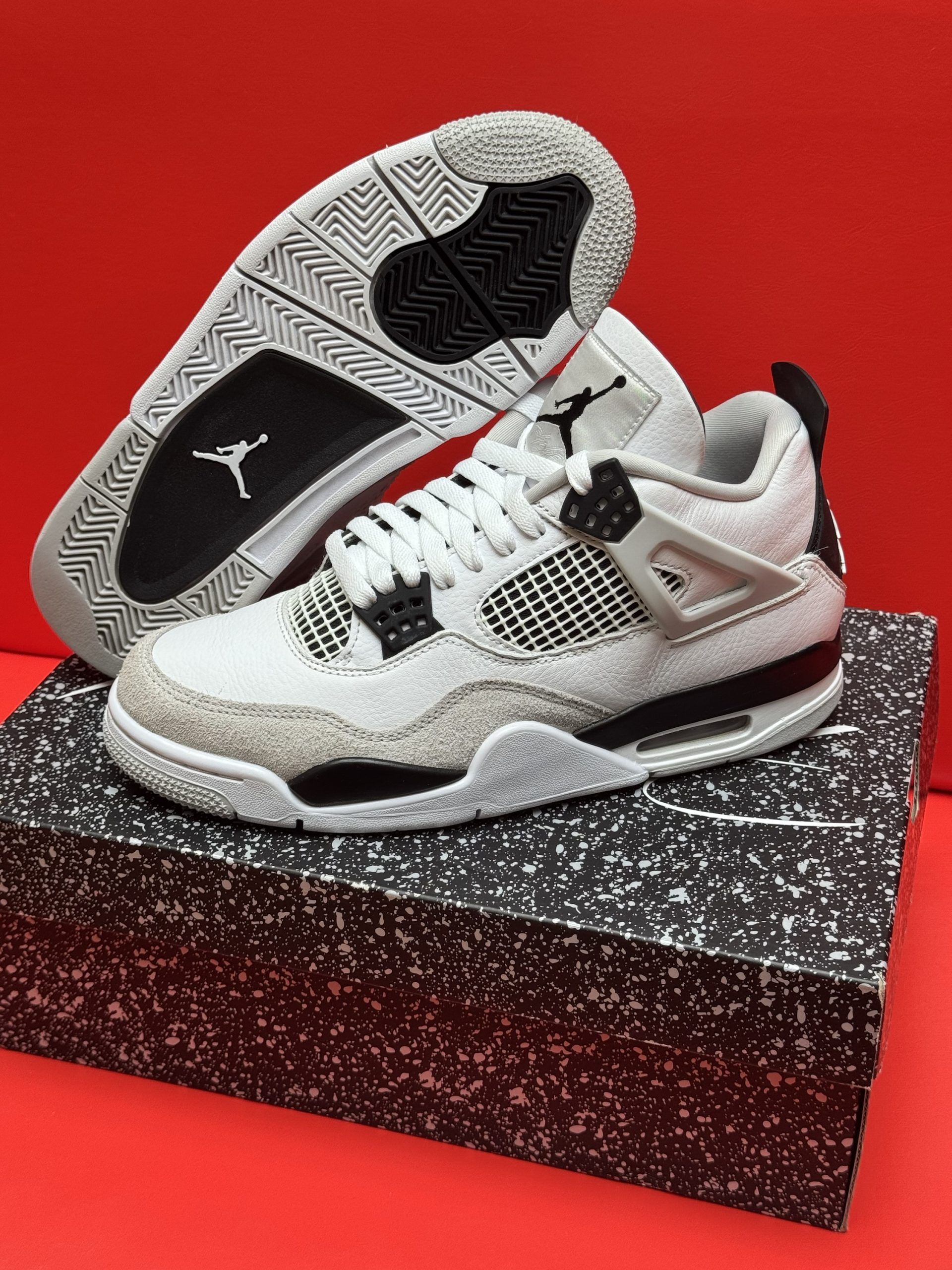 White and gray Air Jordan 4 sneakers on a speckled shoebox, with a red background.