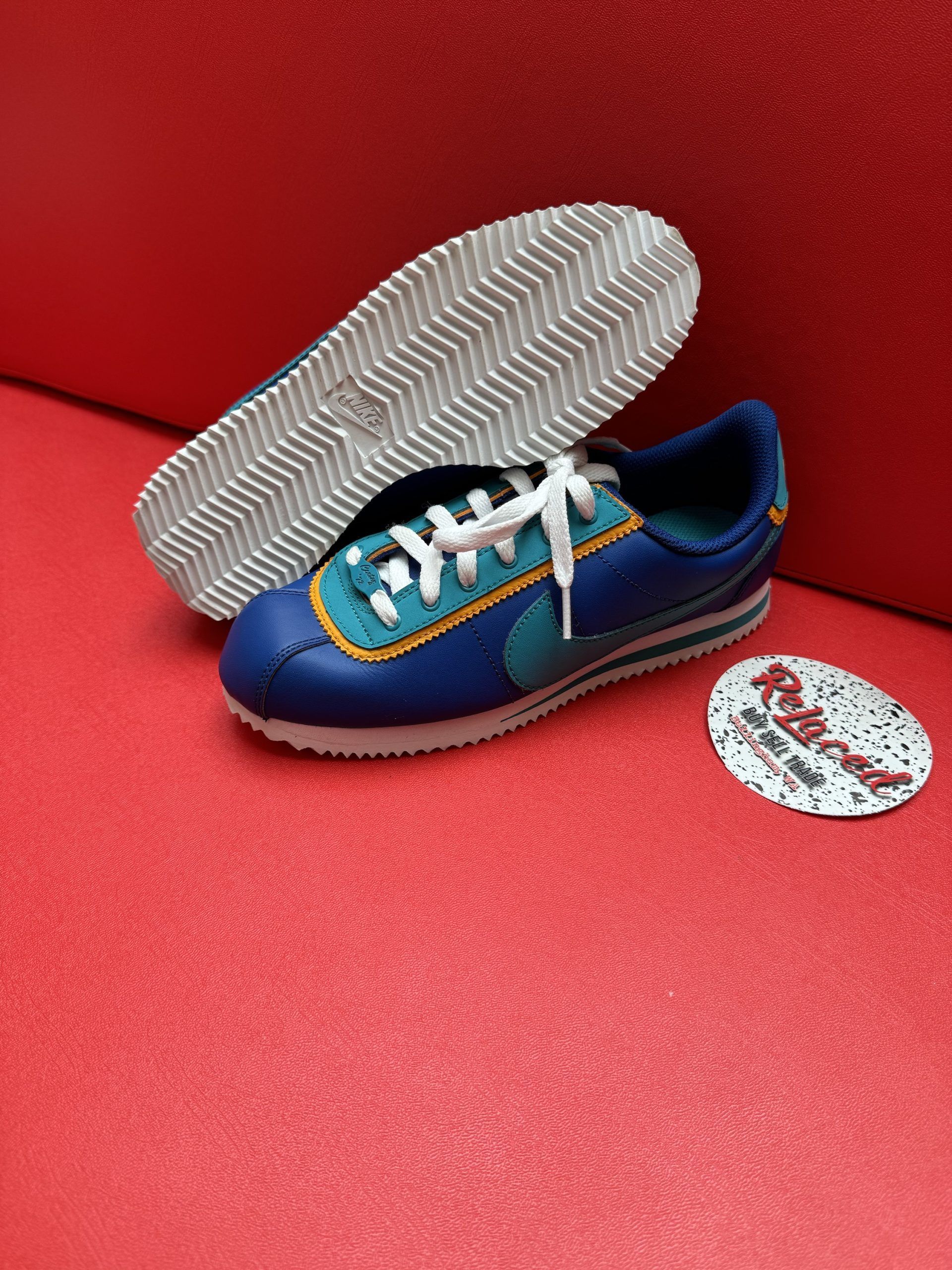 Blue and teal Nike Cortez sneakers with white soles, laces, and a small decorative disc on red surface.