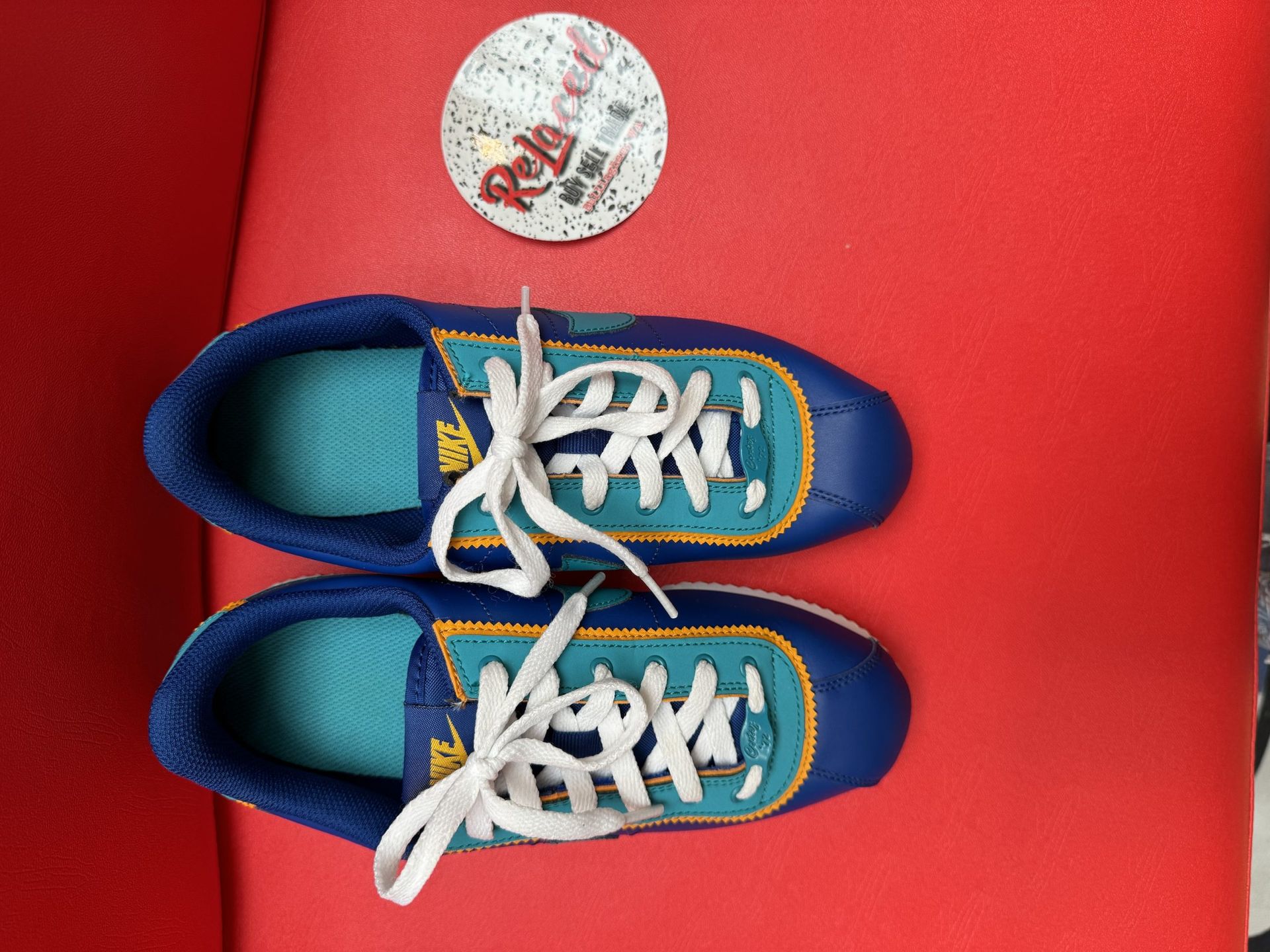Blue and teal Nike Cortez sneakers with white soles, laces, and a small decorative disc on red surface.