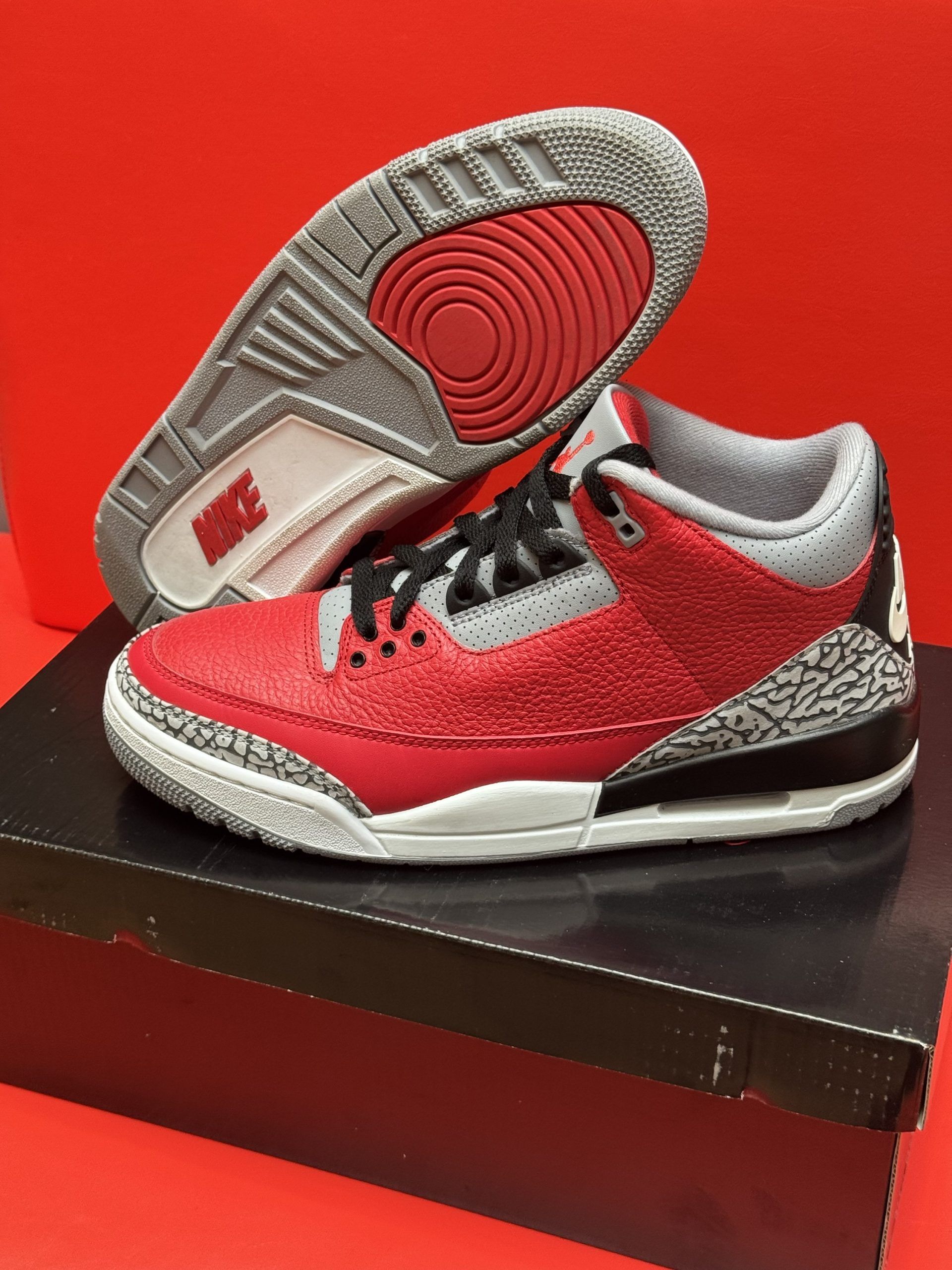 Red and gray Air Jordan 3 sneakers on a black box, with a red background.