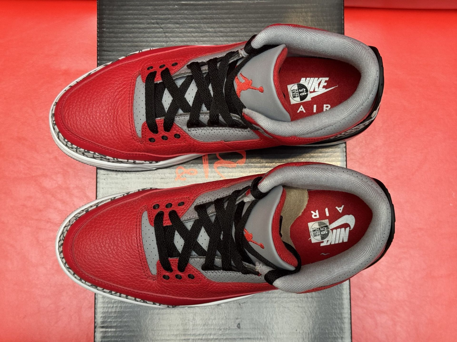 Red and gray Air Jordan 3 sneakers on a black box, with a red background.