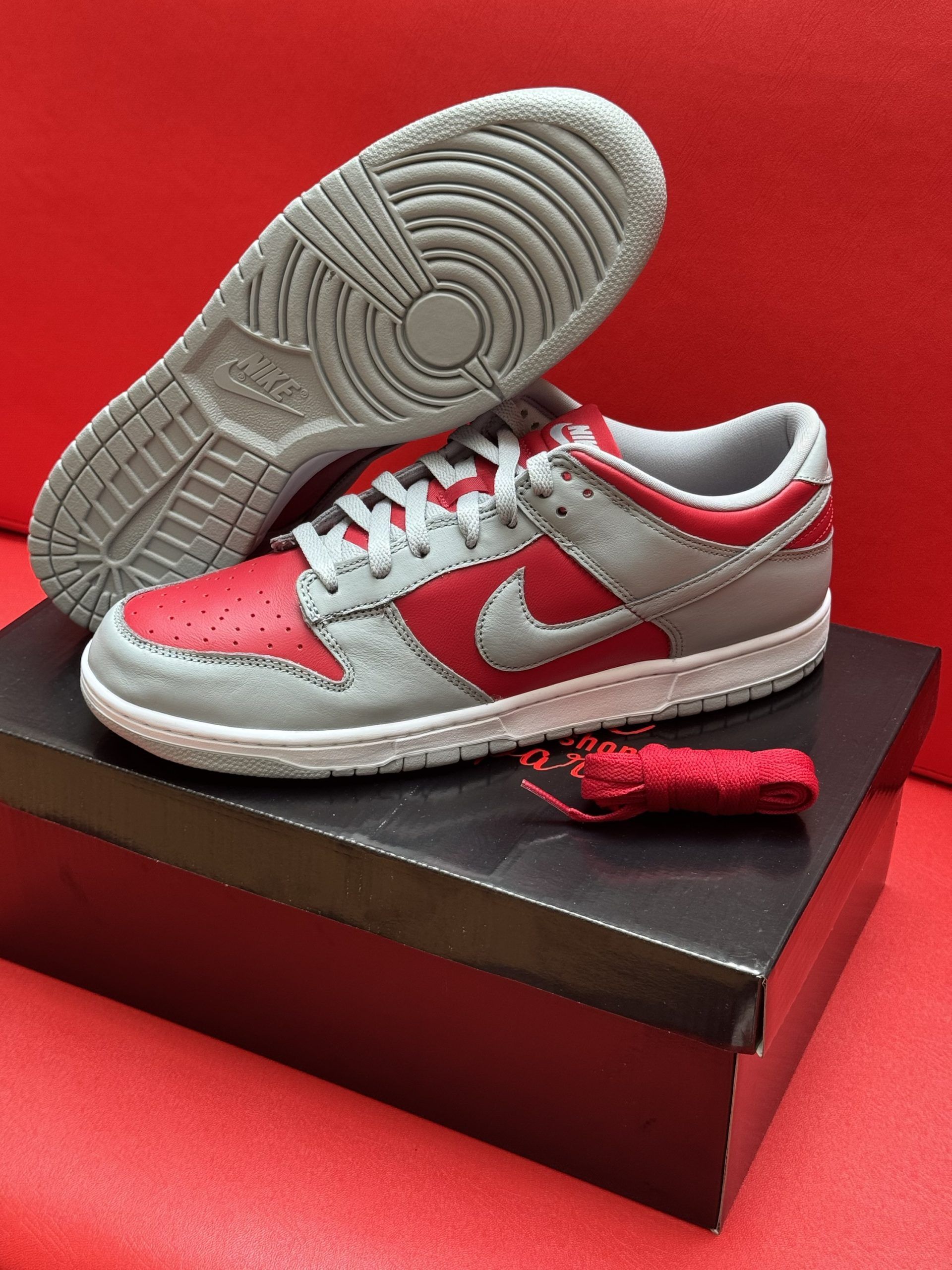 Nike Dunk Low sneakers, red and gray, with gray sole. One shoe rests on its side, the other inverted. Red background.