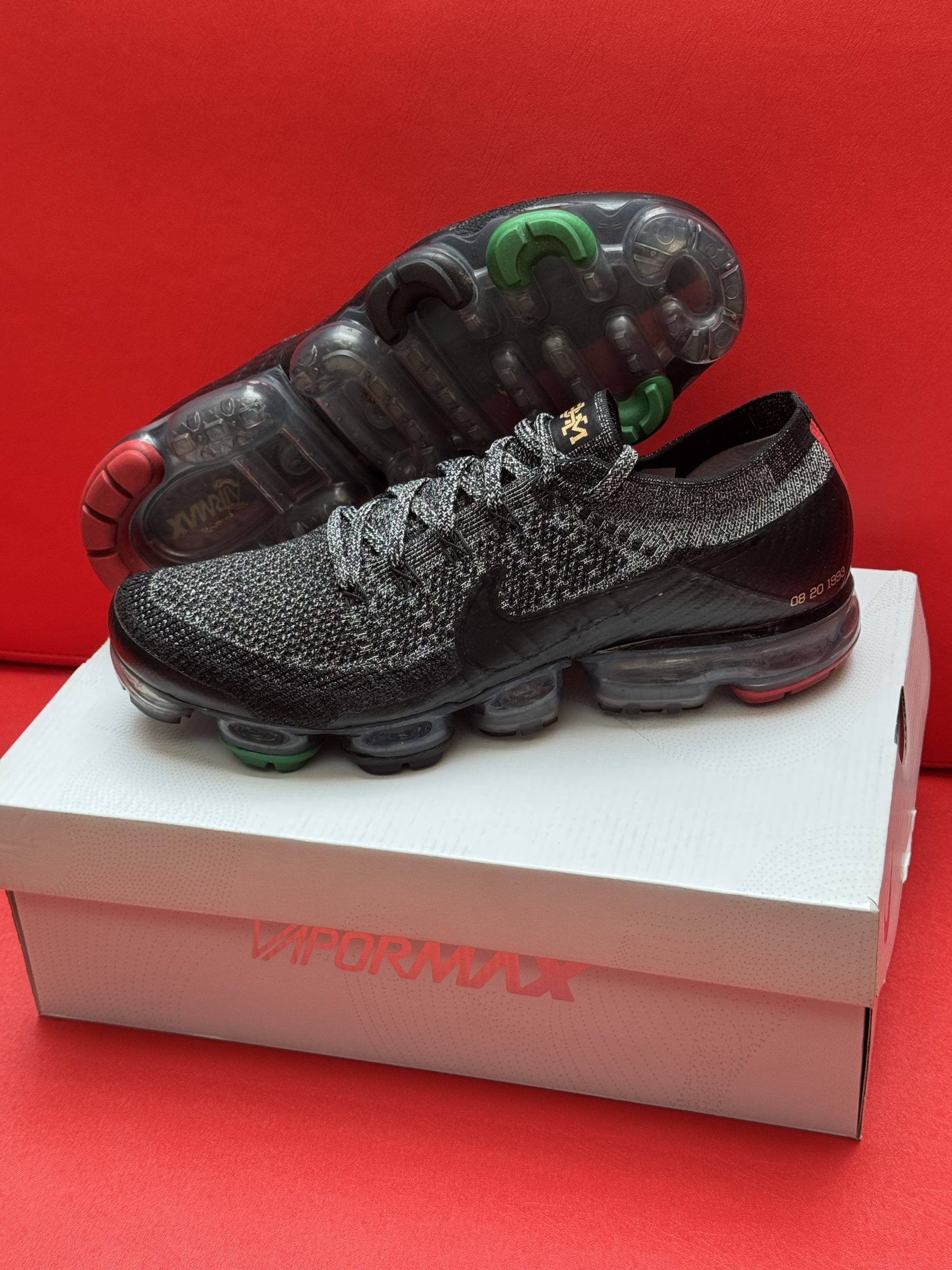 Black and gray Nike Air VaporMax Flyknit sneakers on a white box, red background.