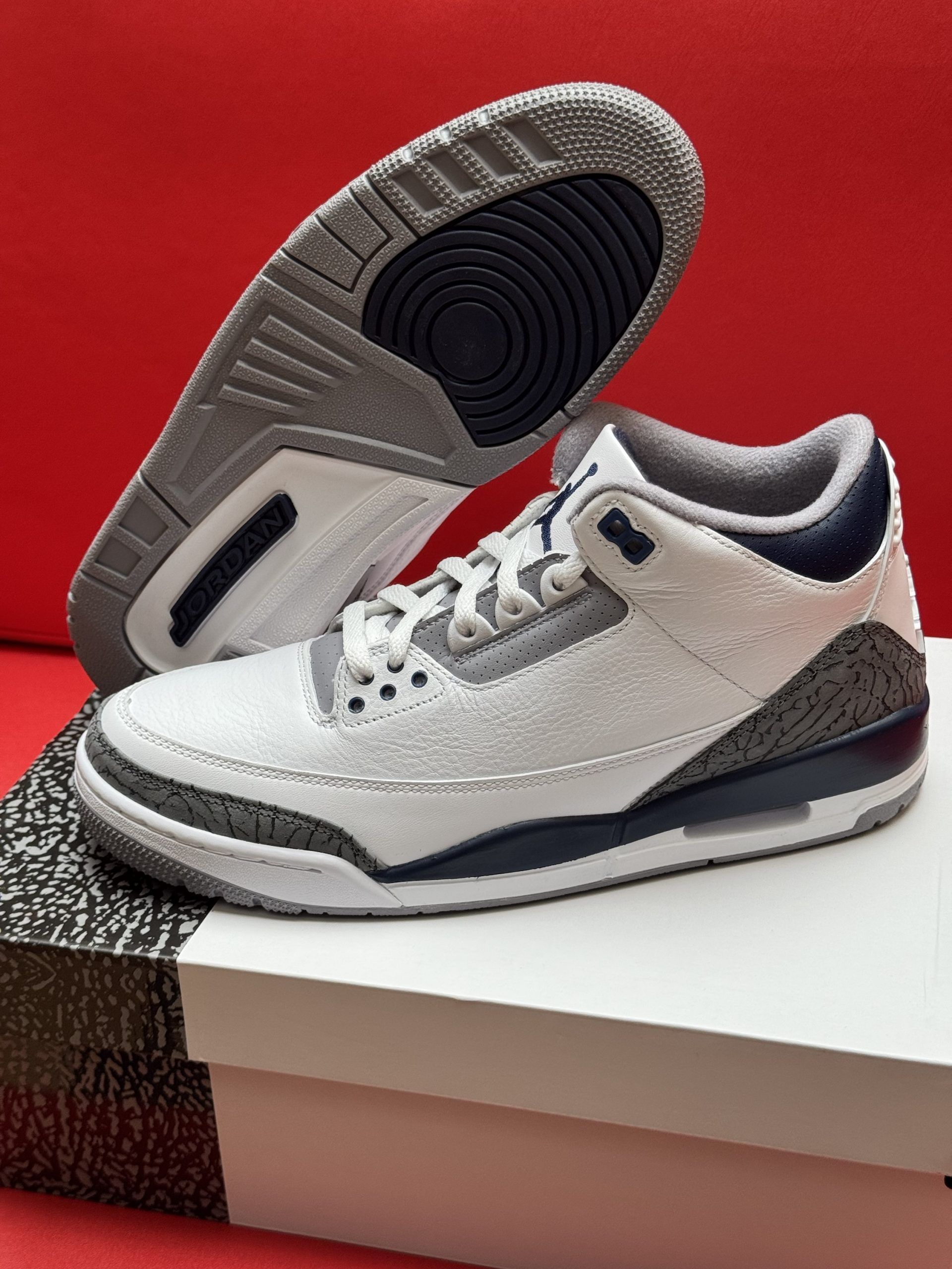 White, gray, and navy Air Jordan 3 sneakers on a box, sole exposed, on a red background.