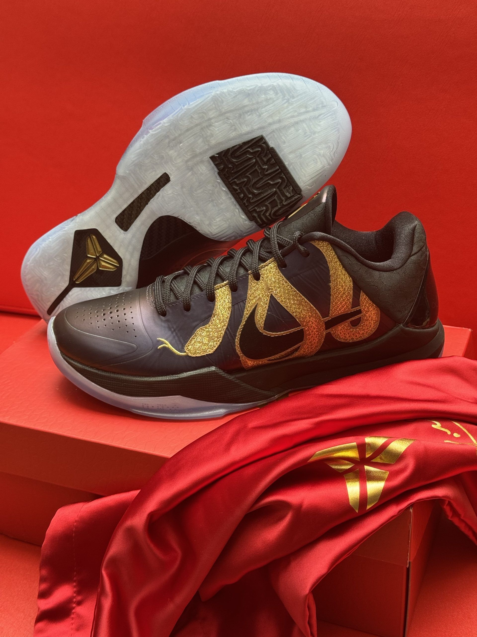 Black and gold Nike Kobe sneakers with a snake design, on a red box with a red cloth, a red background.